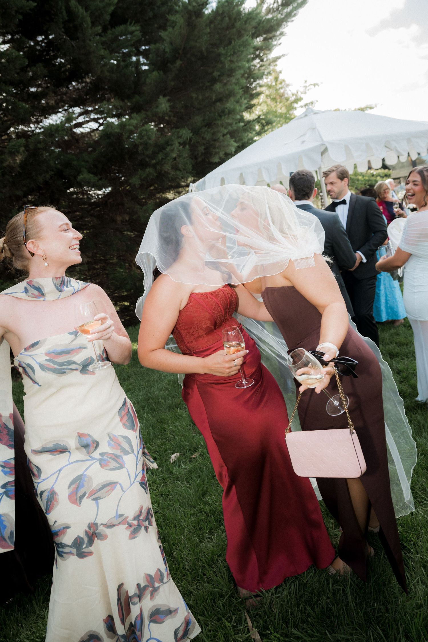 Bride laughing with guests as her veil blows over her face during an outdoor wedding cocktail hour.