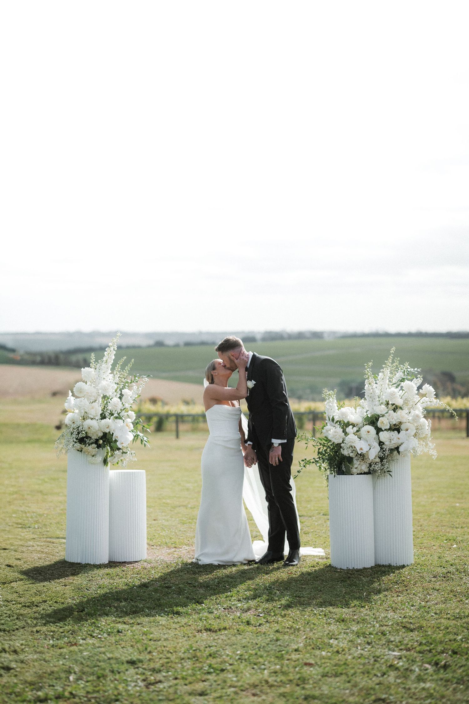 White floral arrangements in milk jugs decorate an outdoor wedding ceremony space on grassy hillside.