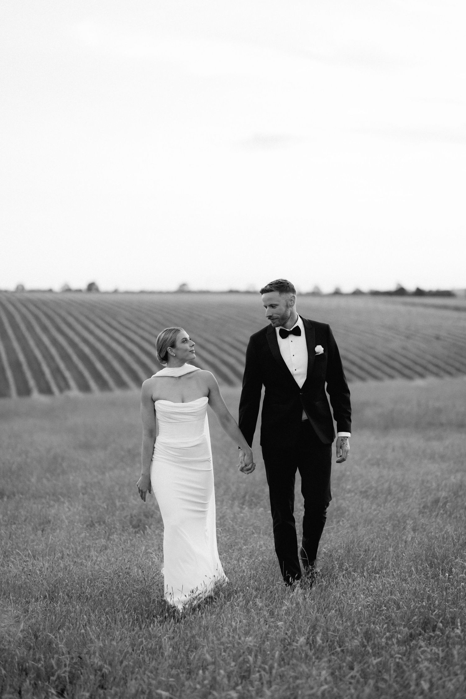 A couple walks together through a scenic field with rows of crops in the background.