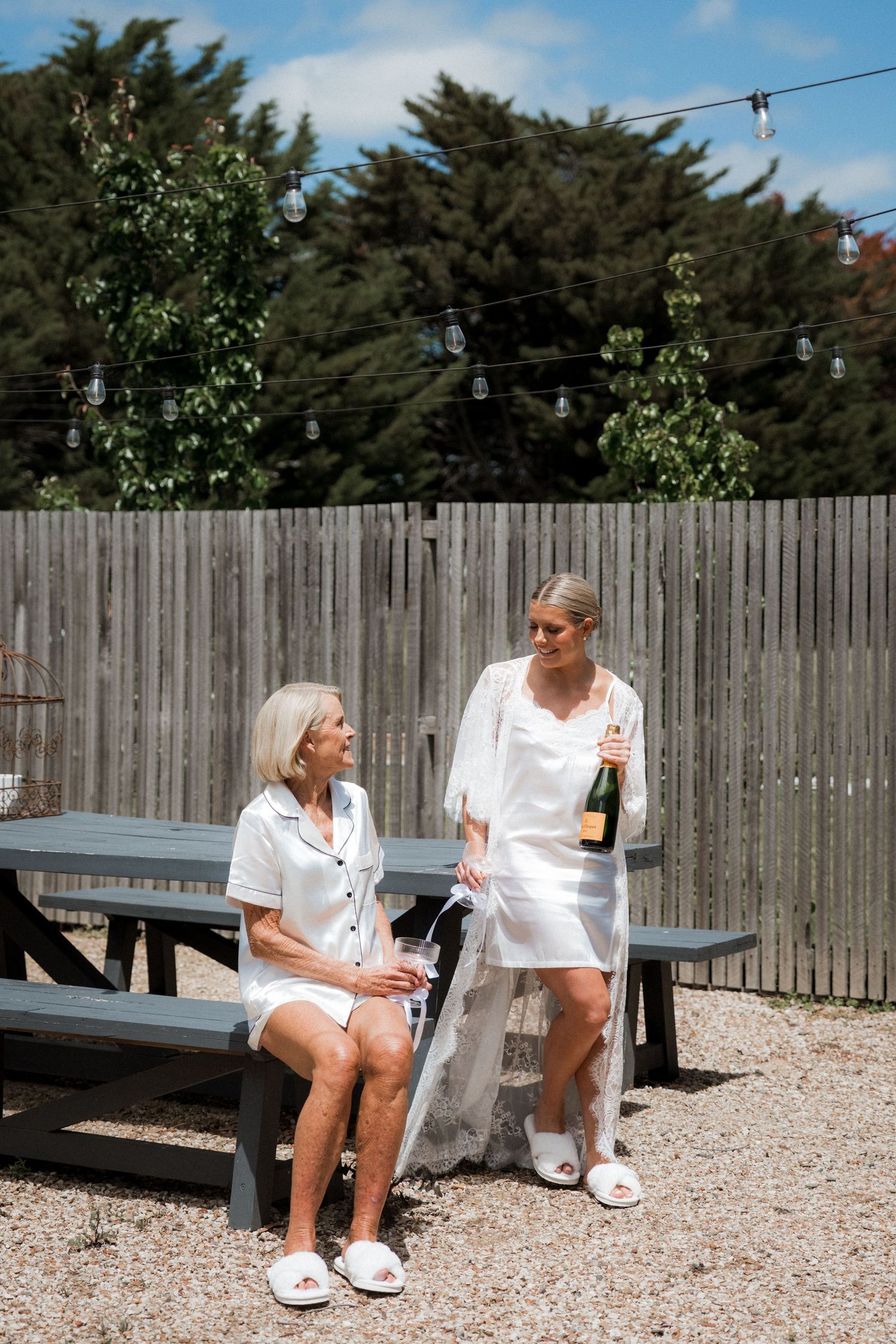 Two people in white outfits sit on outdoor furniture having a casual conversation on a gravel patio.