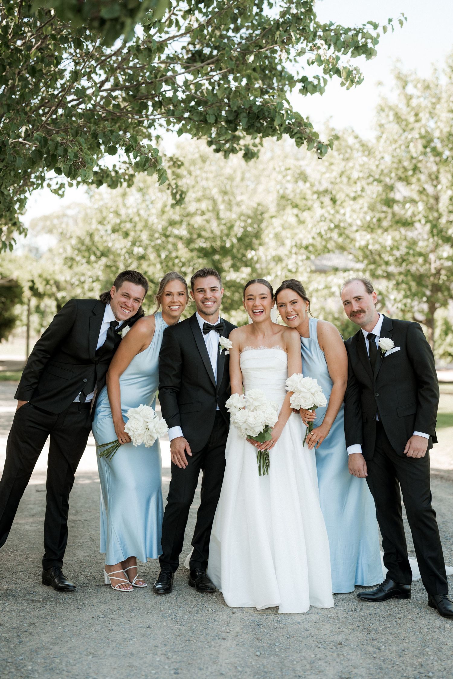 Wedding party poses together outdoors wearing light blue bridesmaid dresses and black tuxedos.
