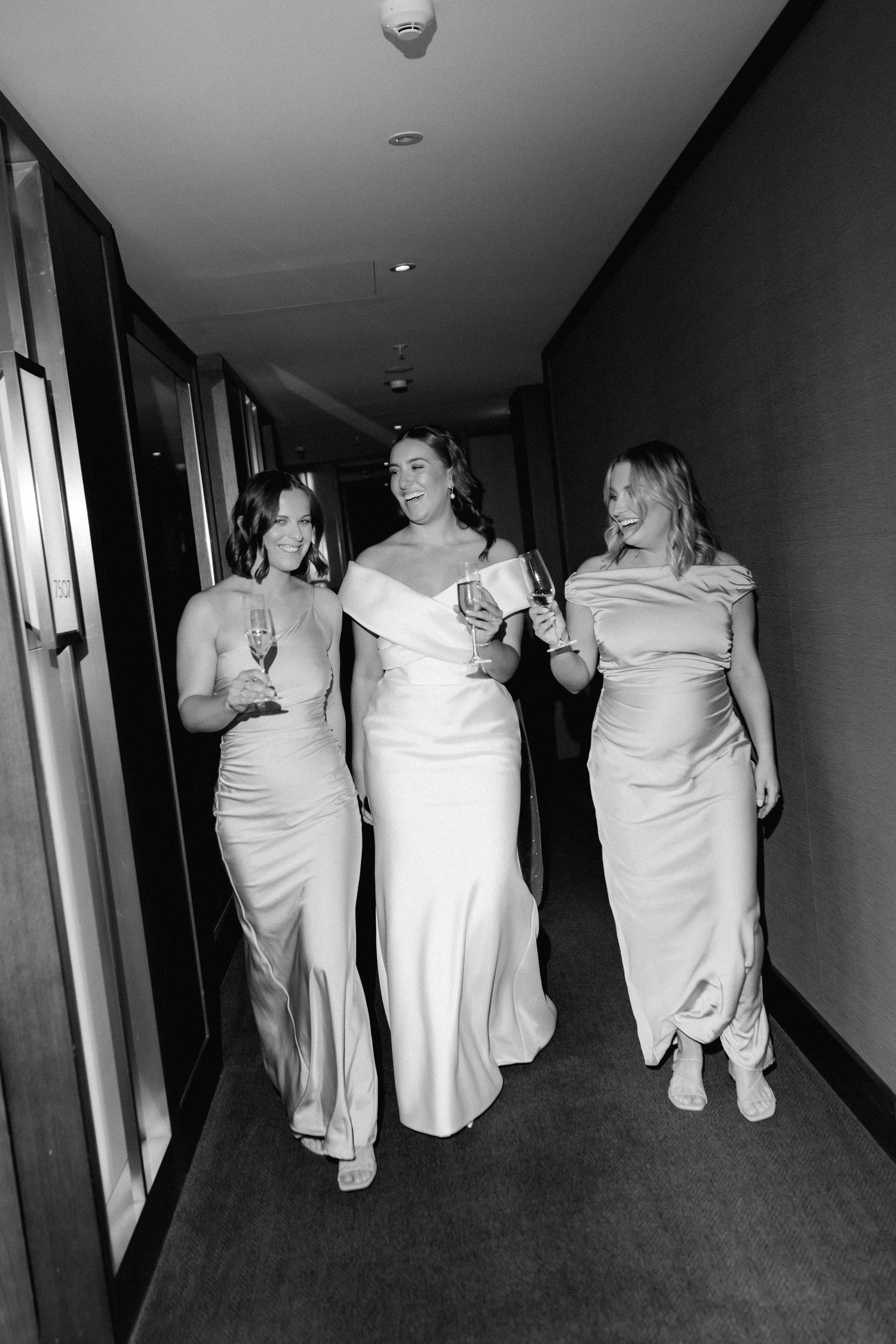 Three bridesmaids in matching light-colored dresses walking down a hallway with champagne glasses.
