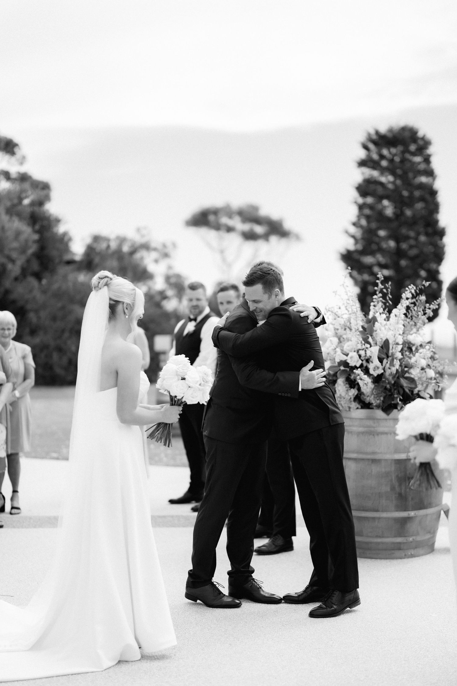 A black and white wedding ceremony moment with couple embracing beside decorative floral barrels outdoors.