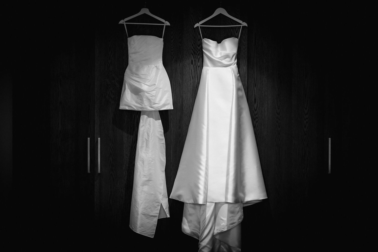 Two white wedding dresses hanging side by side in dramatic black and white contrast.