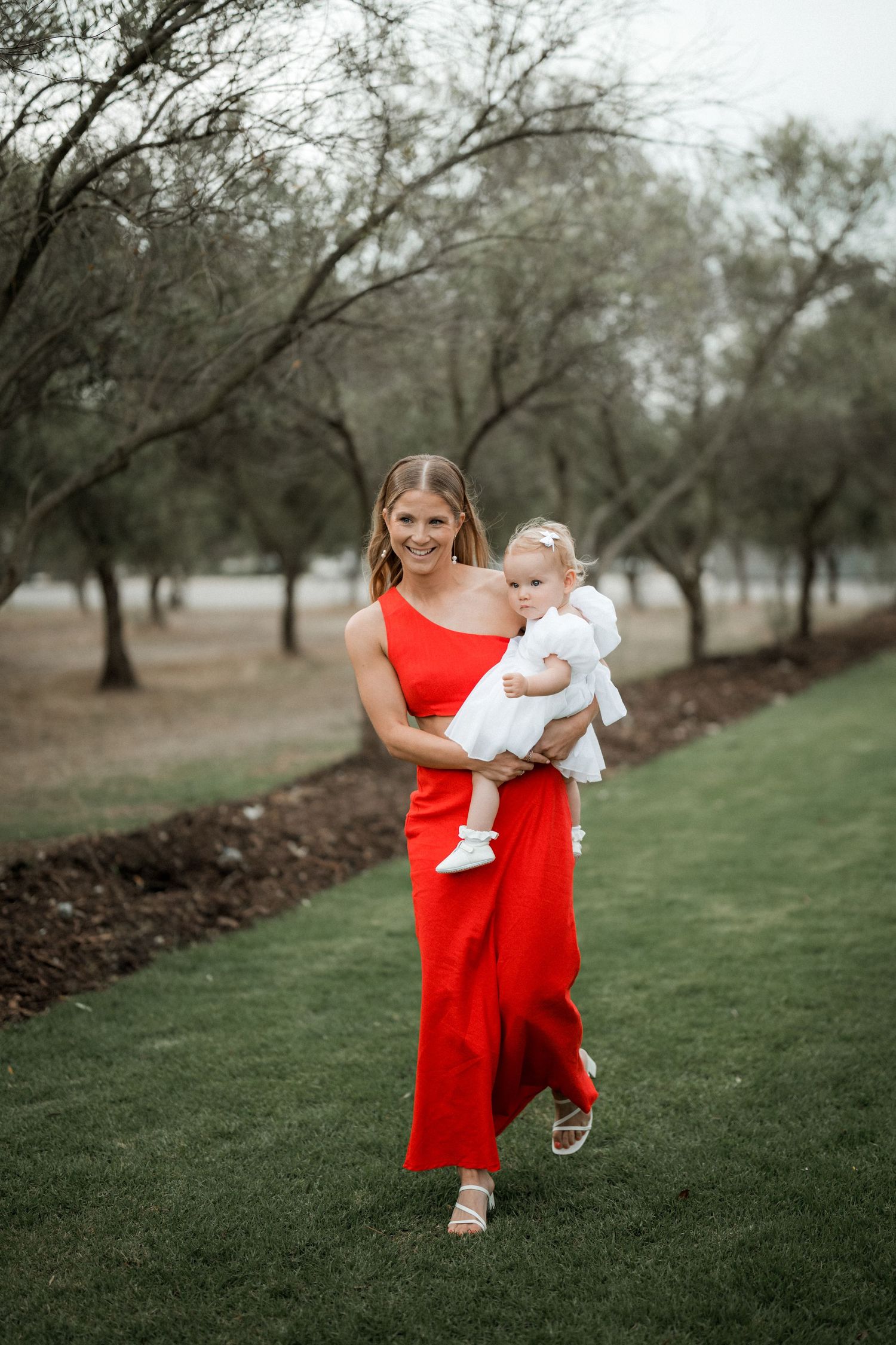 A woman in a flowing red dress holds a baby while walking on manicured grass with trees in the background.