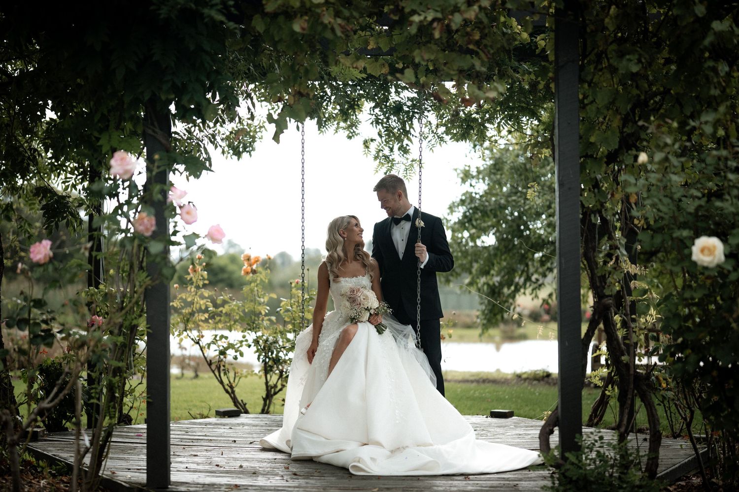 A wedding couple poses under a leafy pergola beside a tranquil lake.
