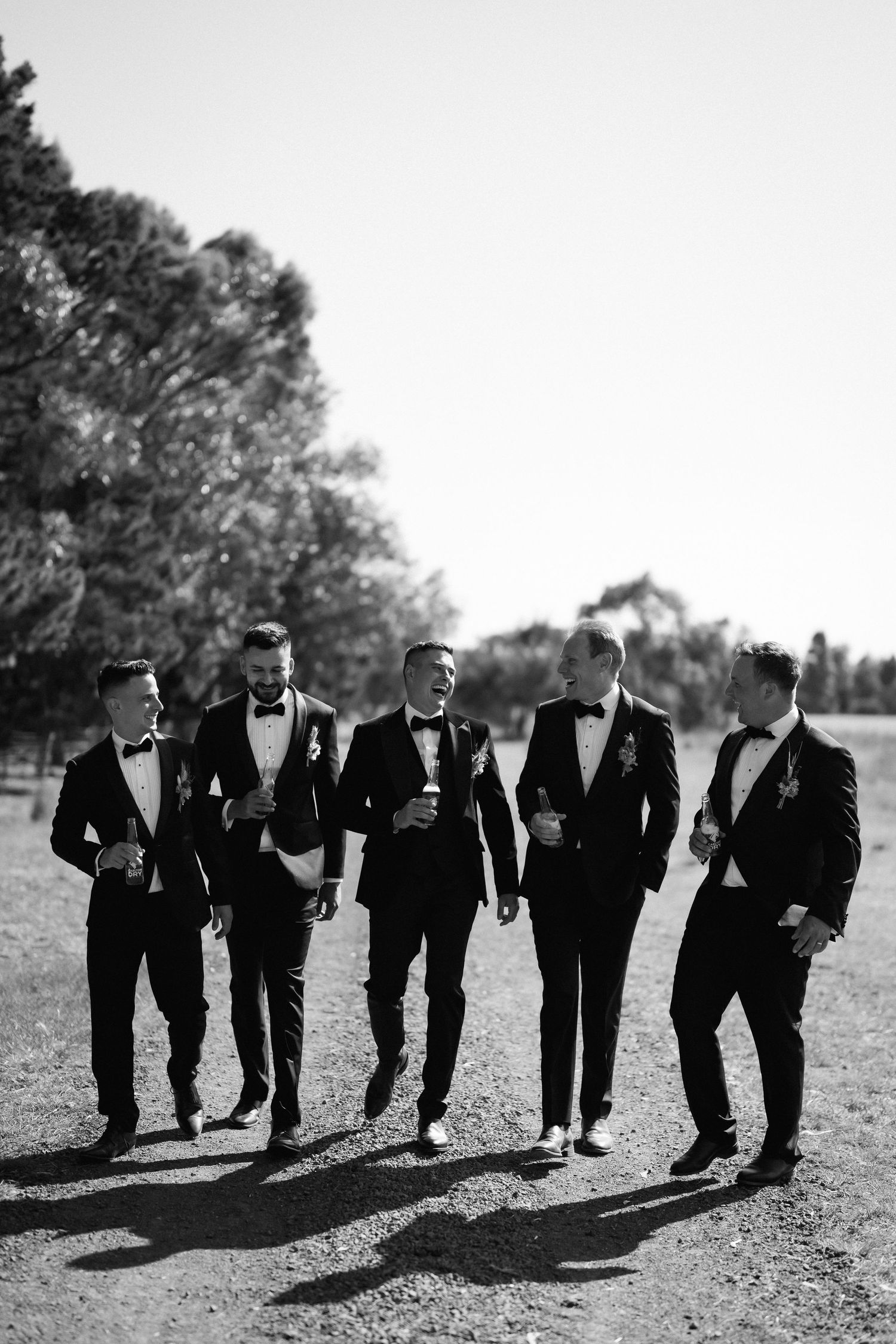 Five groomsmen in black tuxedos walking together on grass in black and white photography.