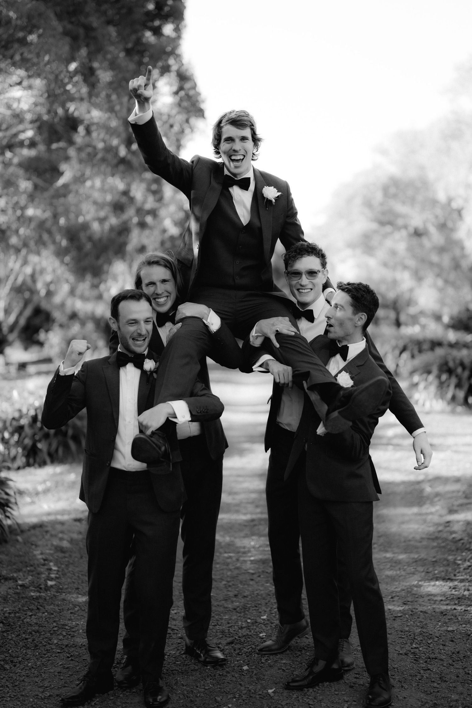 Groomsmen in black tuxedos lift and celebrate during wedding photo session in black and white.