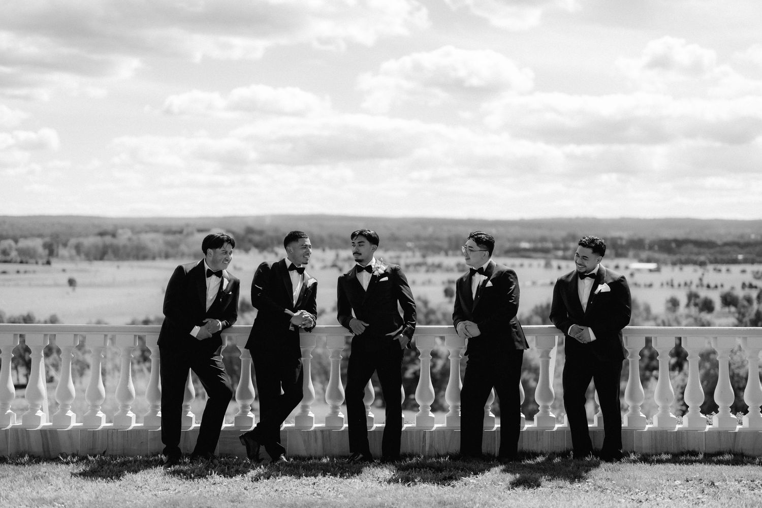 Five groomsmen in black tuxedos and bow ties lean against a stone balustrade overlooking a scenic countryside landscape in black and white.
