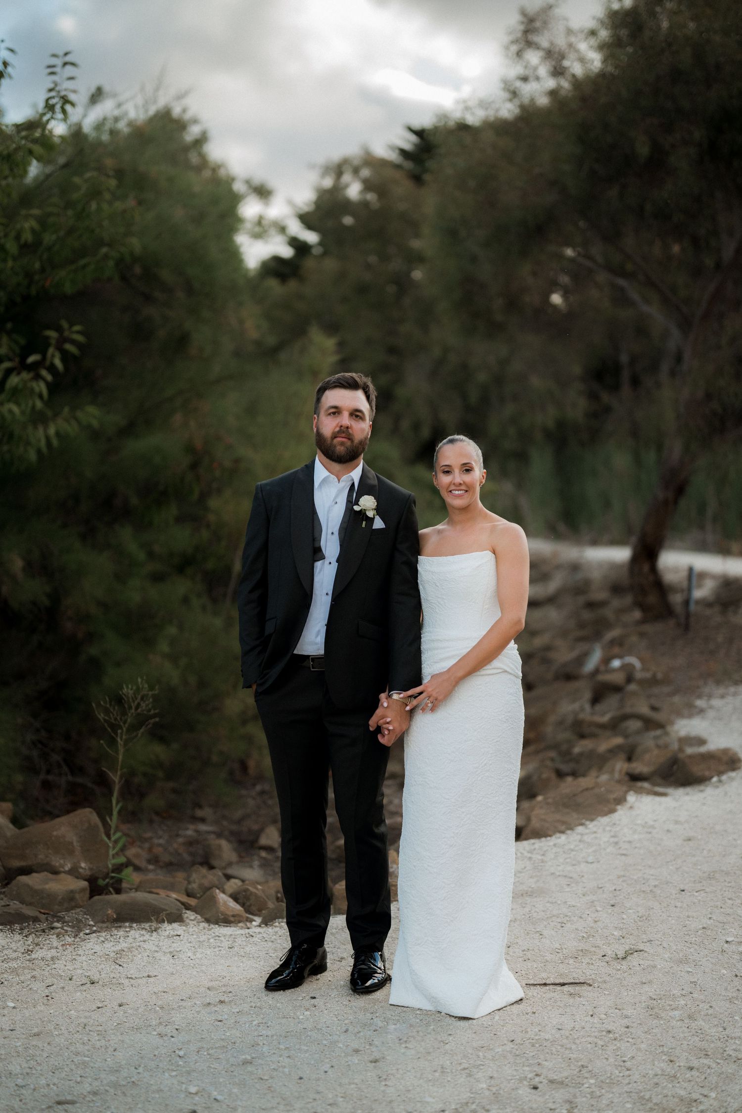 A couple in formal wedding attire poses together on a white sandy beach path surrounded by lush greenery.