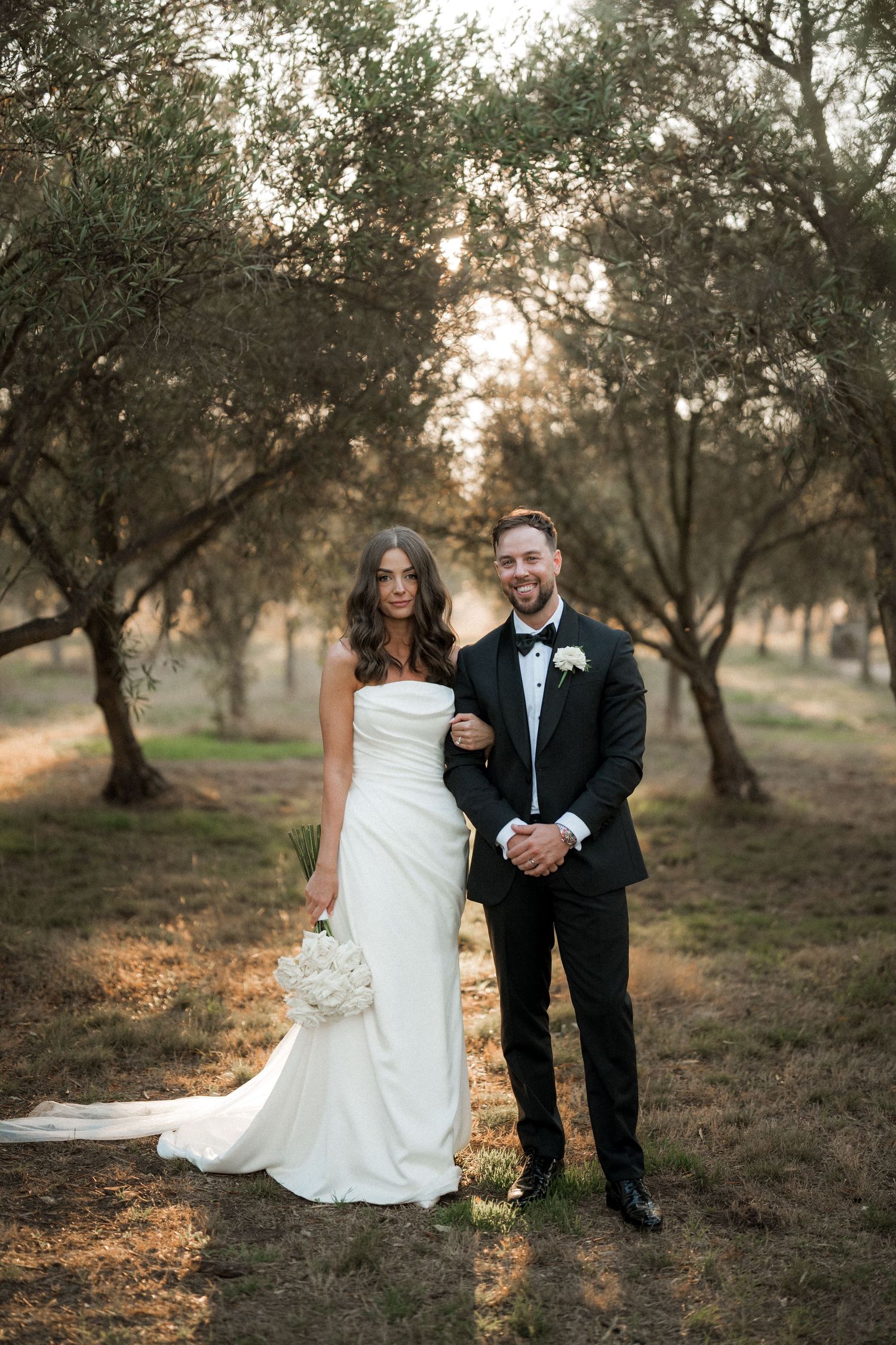 Elegant wedding portrait of couple standing among olive trees at sunset.