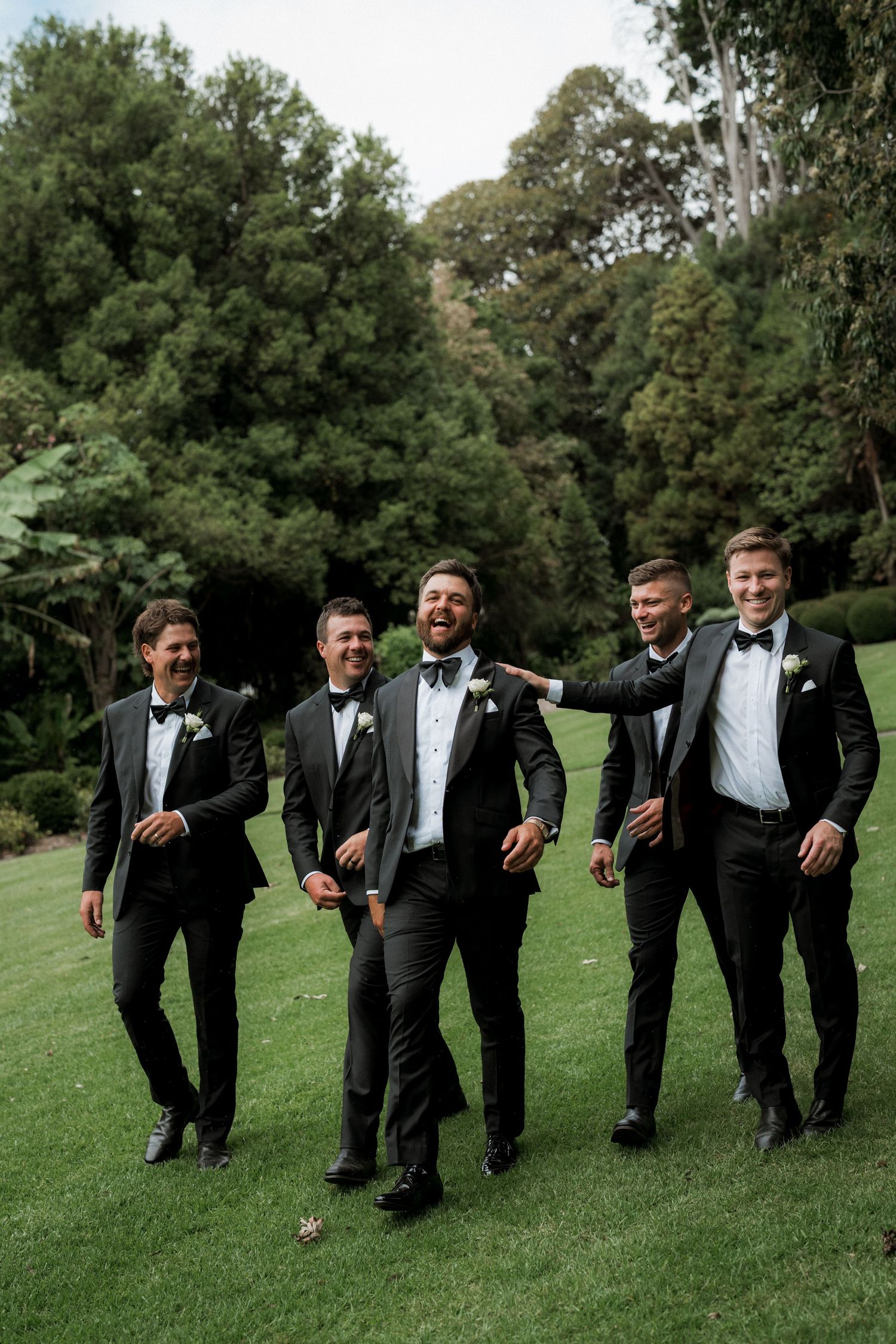 Five groomsmen in black tuxedos and bow ties laughing and walking together on a lush green lawn.