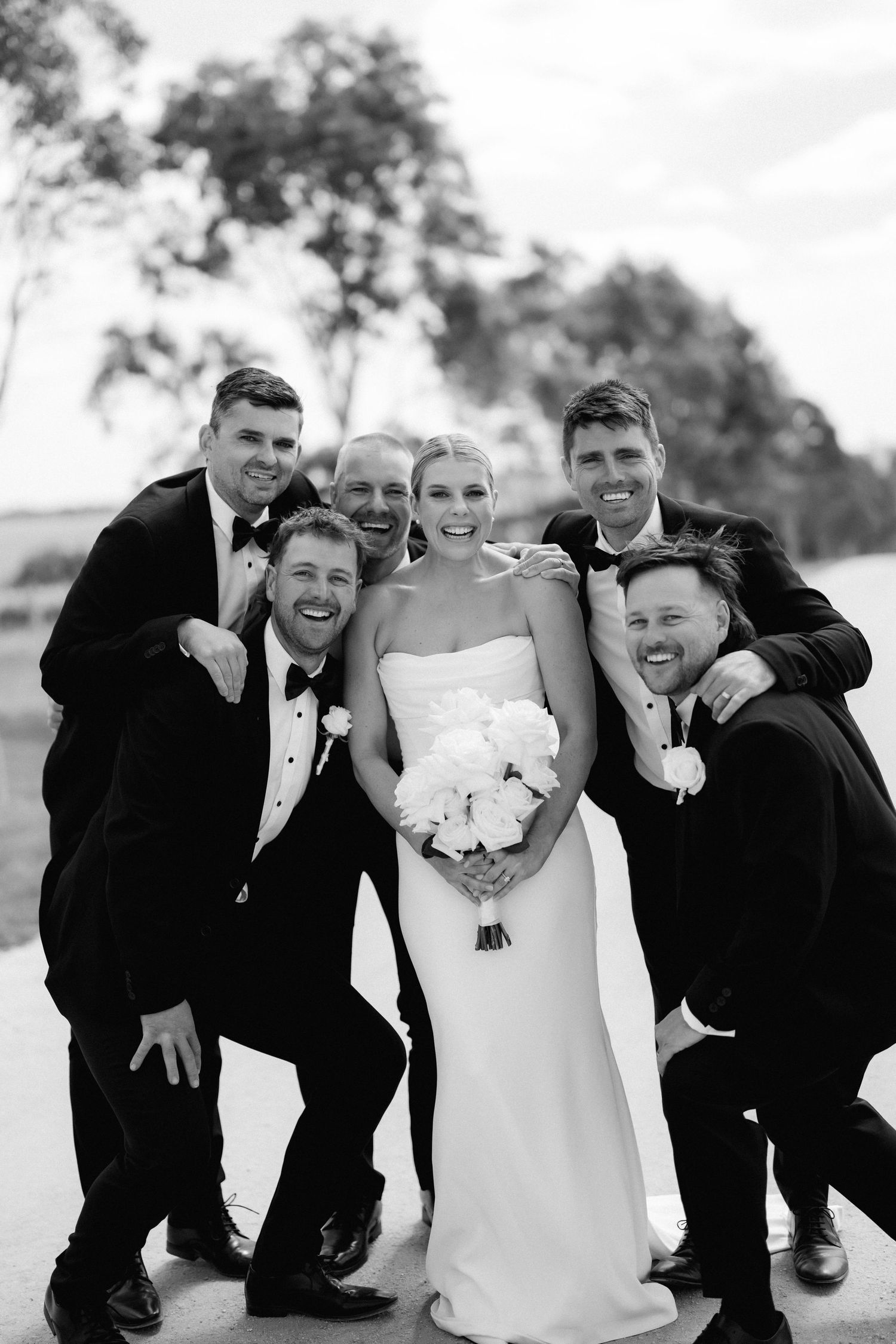 Wedding party in formal attire poses playfully for black and white portrait outdoors.