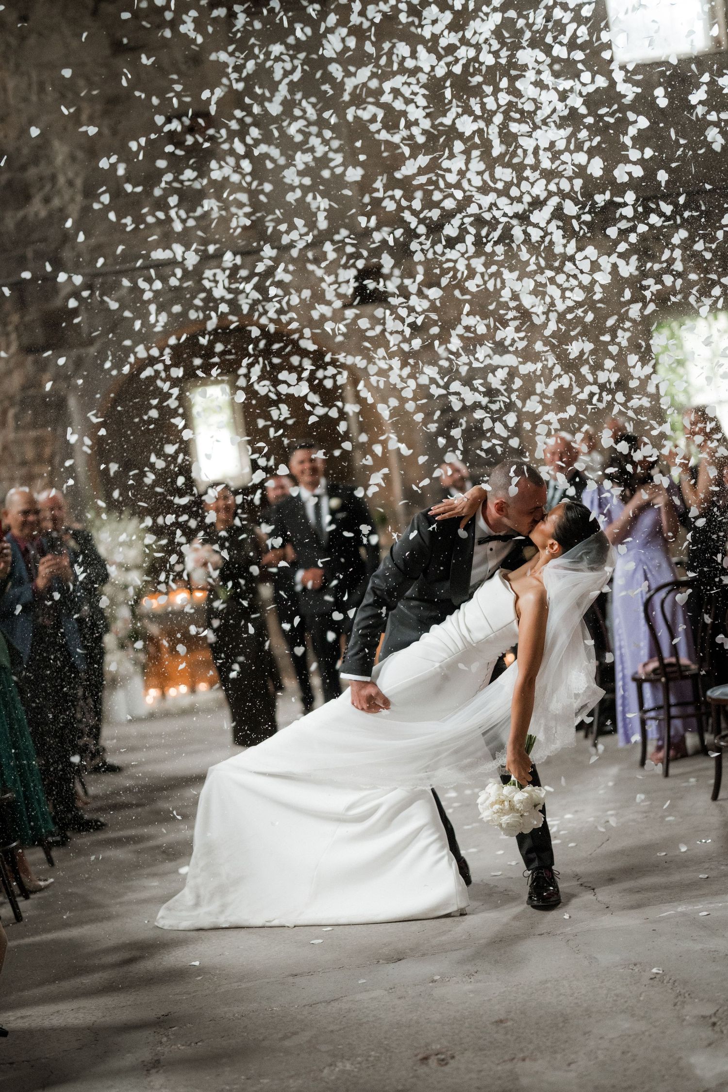 A romantic wedding couple dips and kisses while white flower petals fall around them in a magical outdoor celebration.