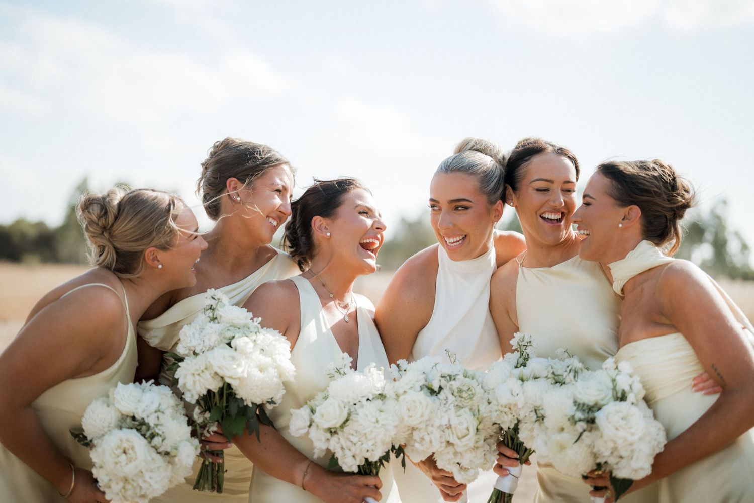 Bridesmaids in light-colored dresses holding white bouquets share a candid laughing moment.