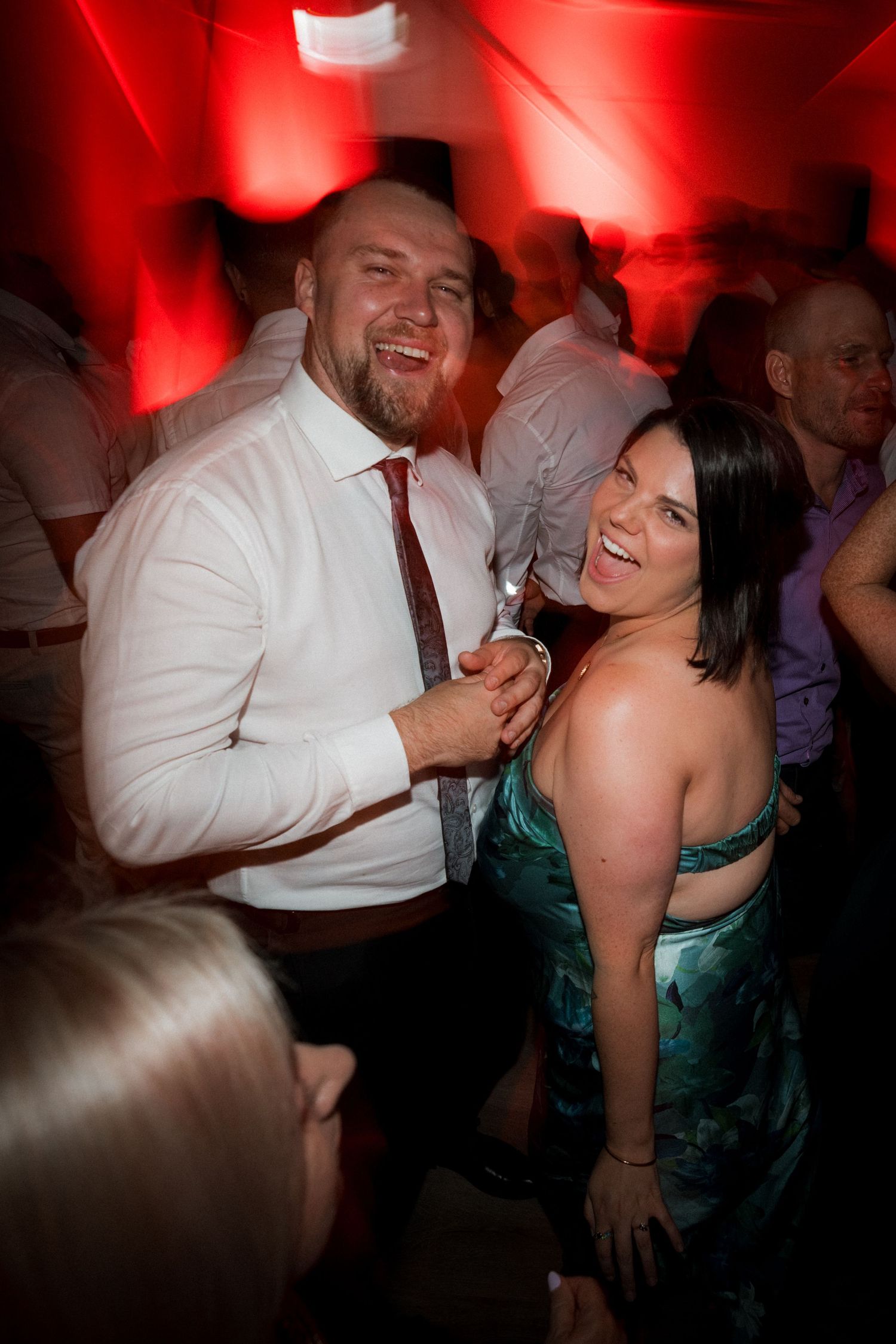 A couple shares a joyful moment dancing together at a party with red lighting in the background.