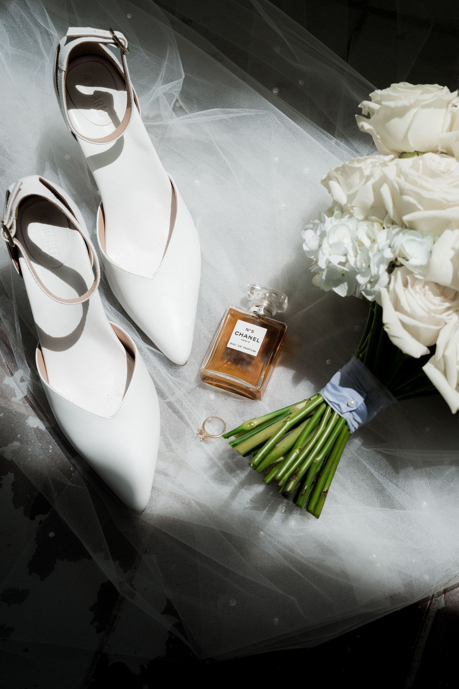 Wedding accessories including white shoes, perfume bottle and floral bouquet arranged on tulle fabric.