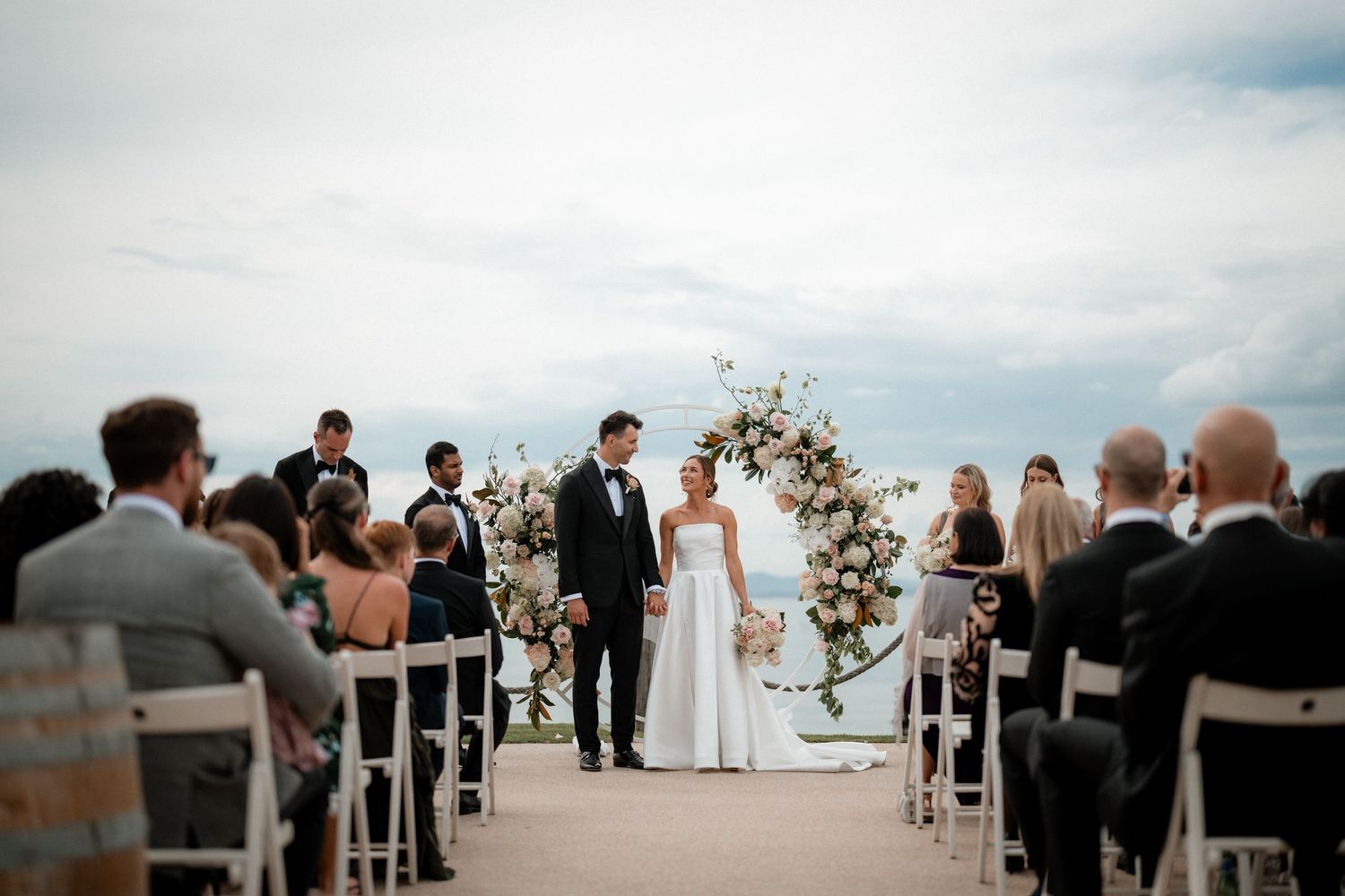 An outdoor wedding ceremony with guests seated in white chairs facing a floral arch against a cloudy sky.