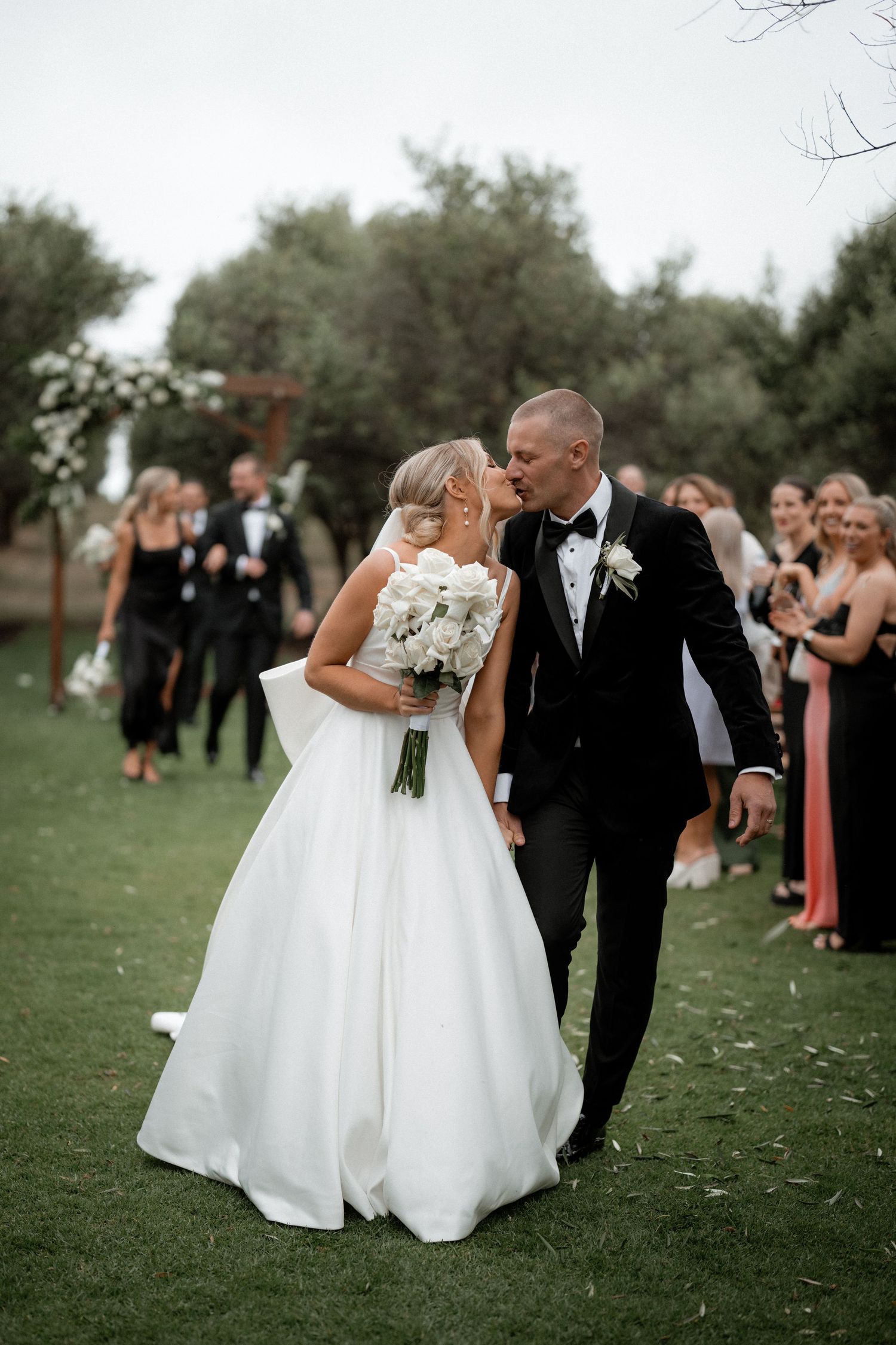 A newlywed couple walking on grass surrounded by wedding guests, bride in white gown holding bouquet.