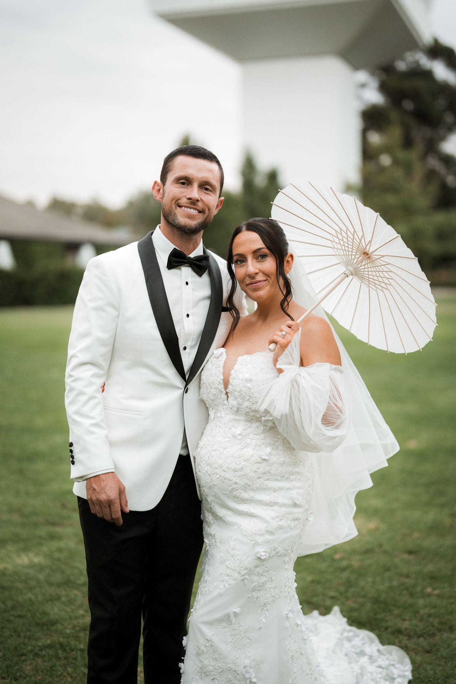 Wedding couple poses outdoors with a white parasol and formal attire.