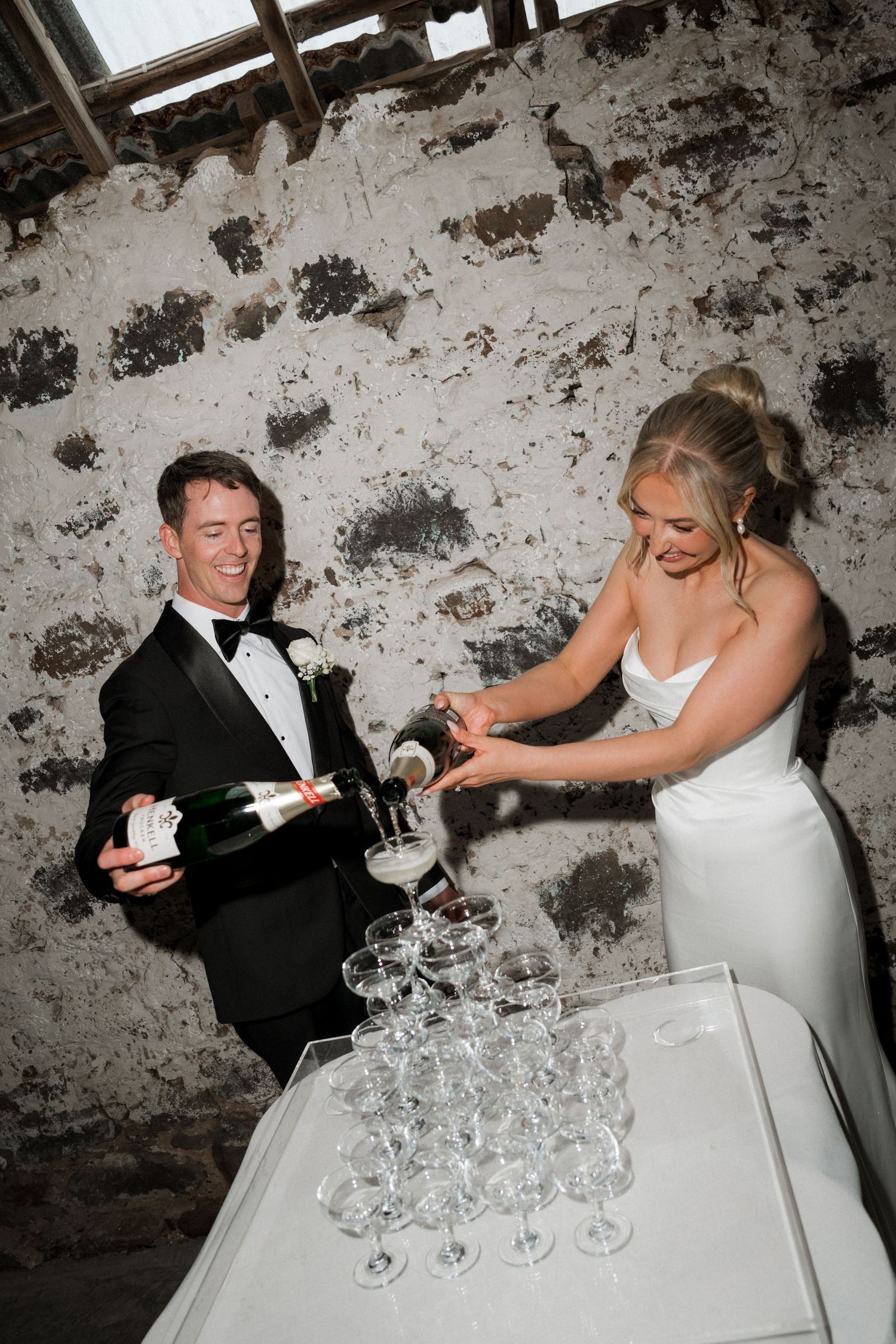 Wedding couple pouring champagne into tower of glasses against rustic wall.