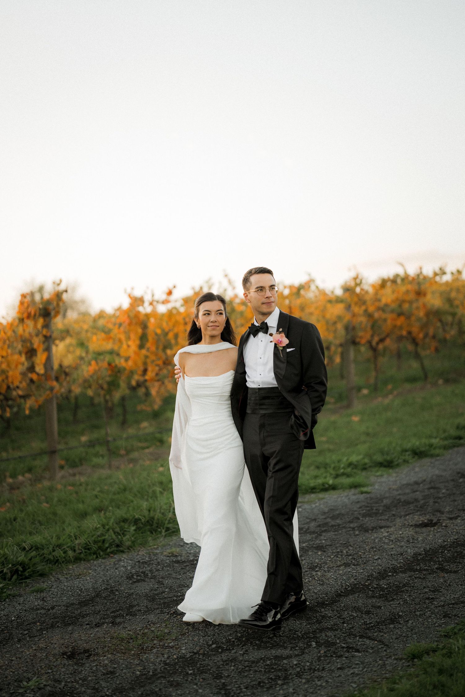 Bride in white strapless gown and groom in black tuxedo pose together in front of golden autumn vineyard.