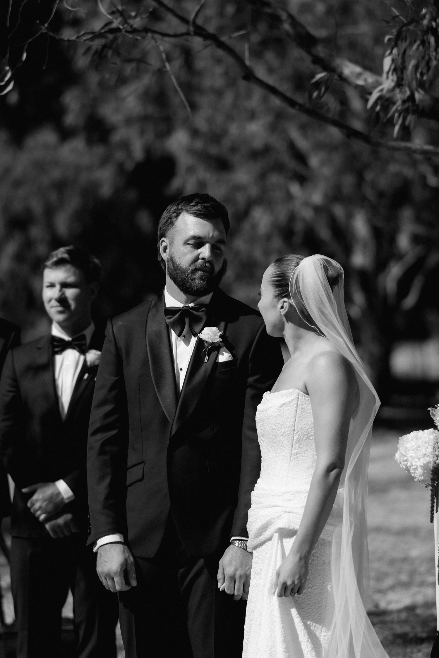 Black and white photo of groom gazing at veiled bride during outdoor wedding ceremony.