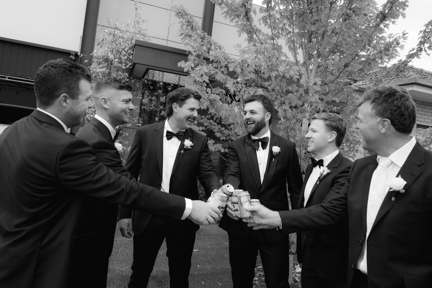 Groomsmen in black tuxedos toasting together outdoors in a black and white wedding photo.