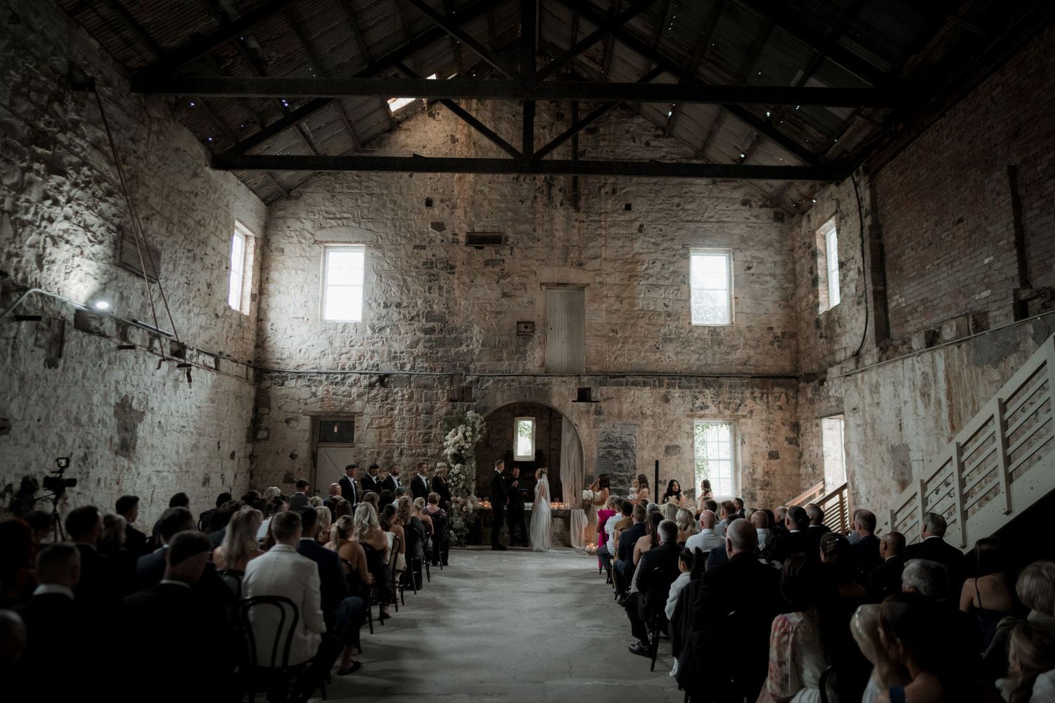 A rustic wedding ceremony takes place inside an old stone barn with exposed brick walls and light streaming through windows.