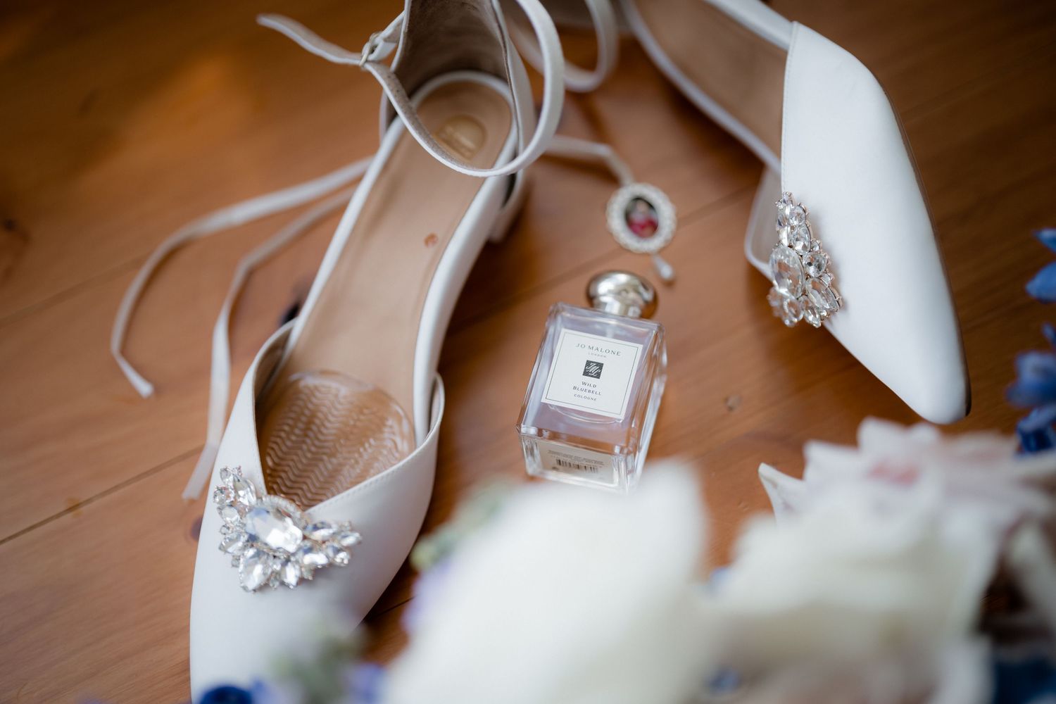 White bridal shoes with lace details arranged with a perfume bottle and ring on wooden surface.