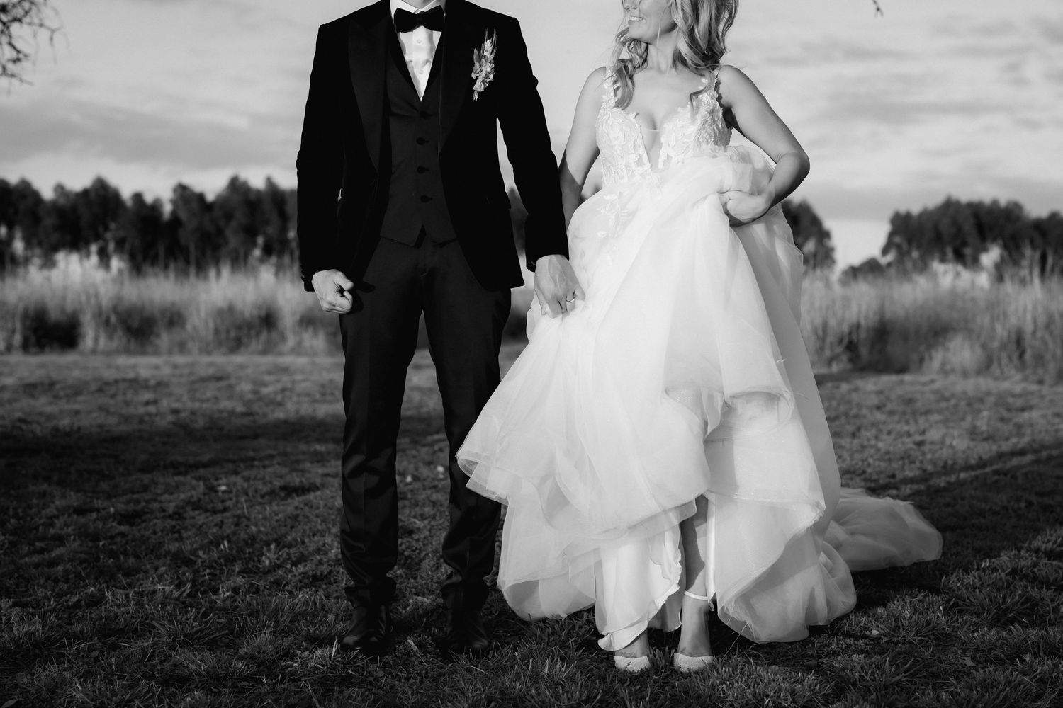 Black and white photo of a bride in flowy wedding dress and groom in tuxedo standing in an outdoor field.