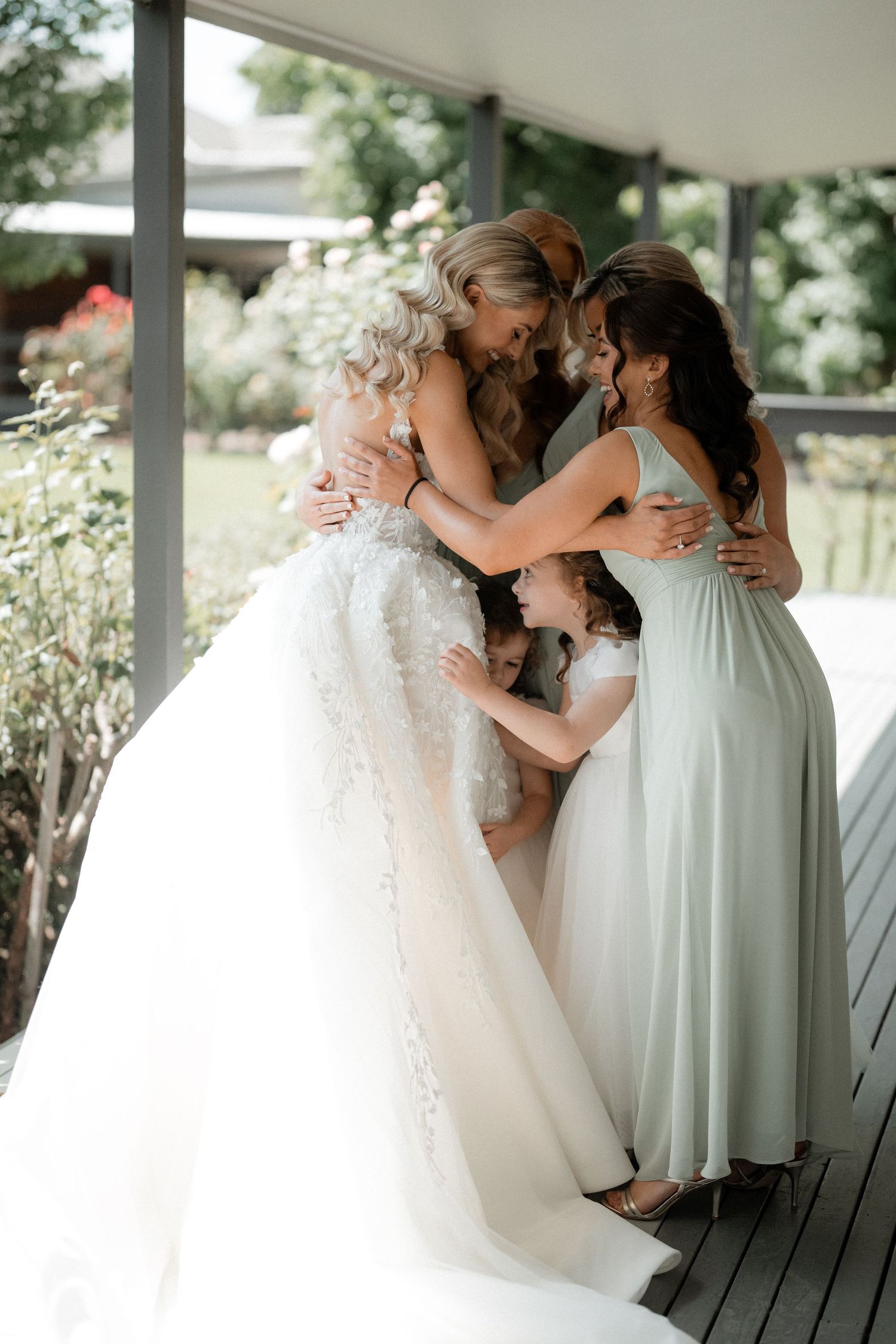 Wedding party embracing in an emotional moment on a covered porch with natural lighting.