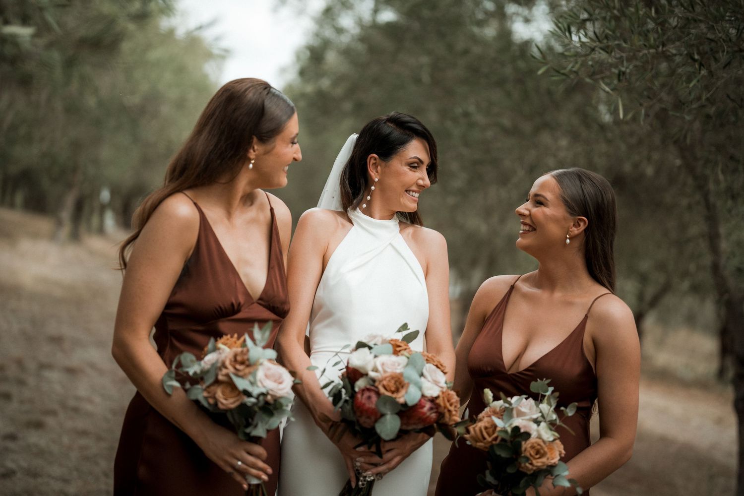 Bride in white dress and bridesmaids in brown dresses share laughter while holding floral bouquets.