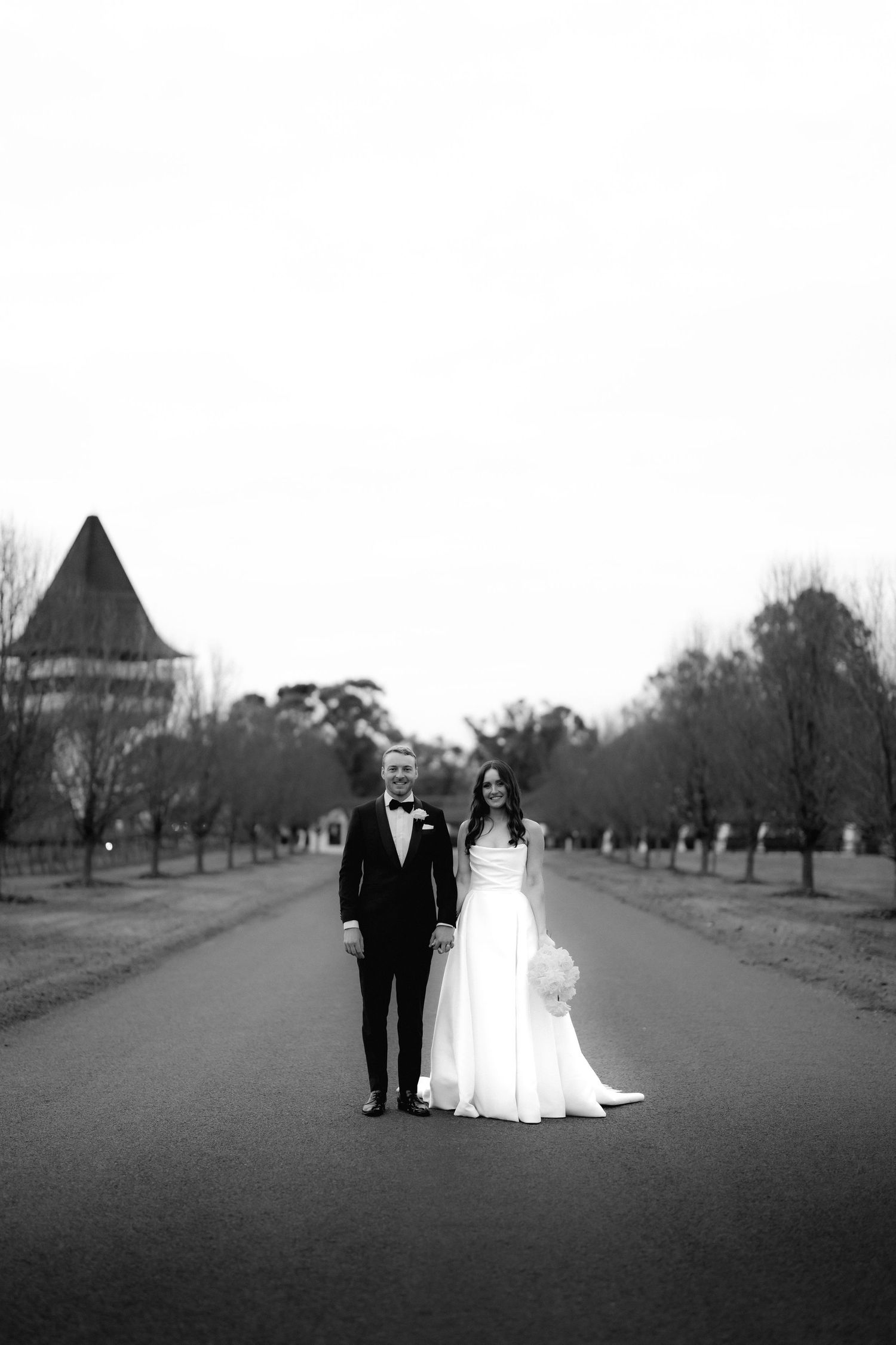 Elegant black and white photo of wedding couple standing on tree-lined path with historic building.