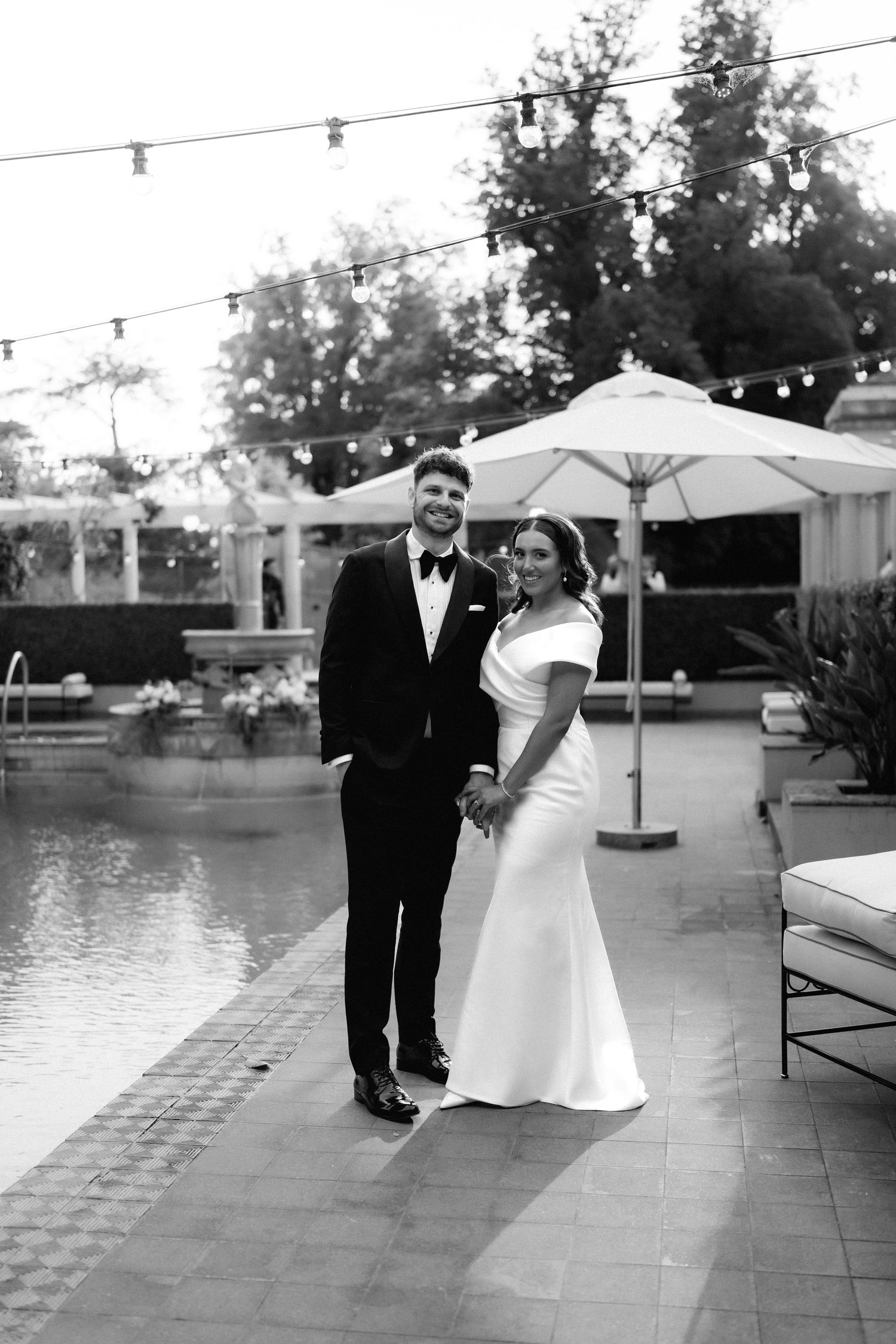 Couple stands by poolside at an outdoor wedding reception with string lights and umbrellas in black and white.