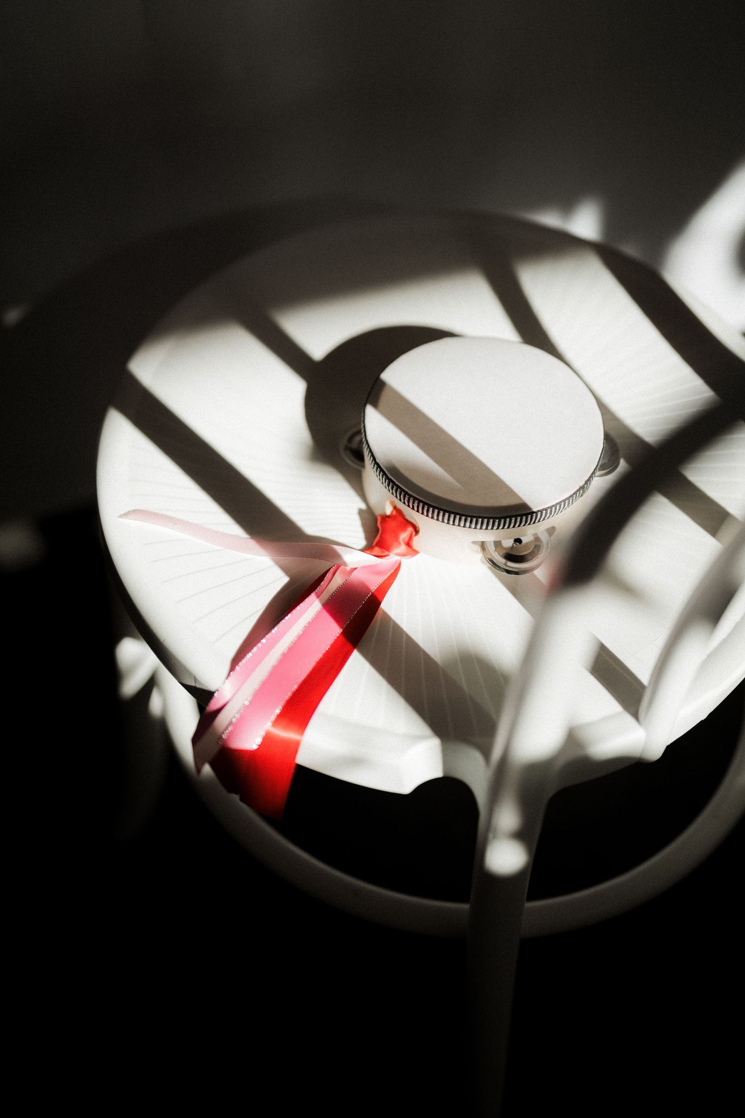A perfume bottle with red ribbon on white stool, dramatic shadows from window light creating geometric patterns.