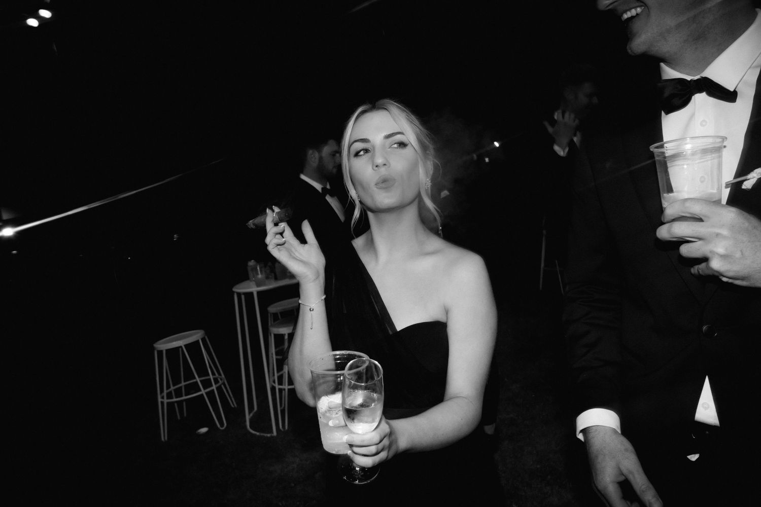 Black and white photo of a woman in an elegant dress holding a drink and cigarette at a formal evening event.