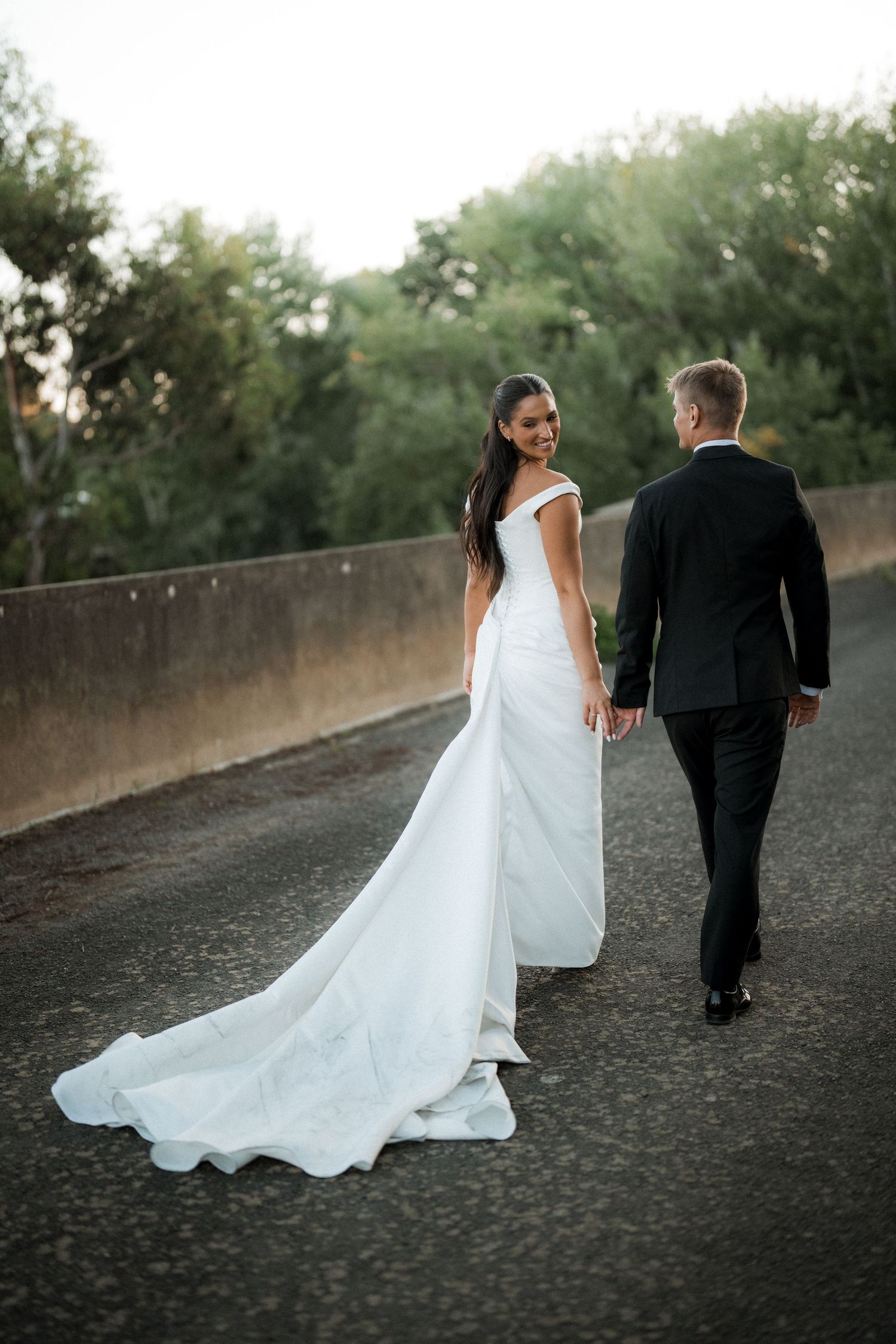 Couple walking along a path as the bride's elegant white dress train trails behind them.