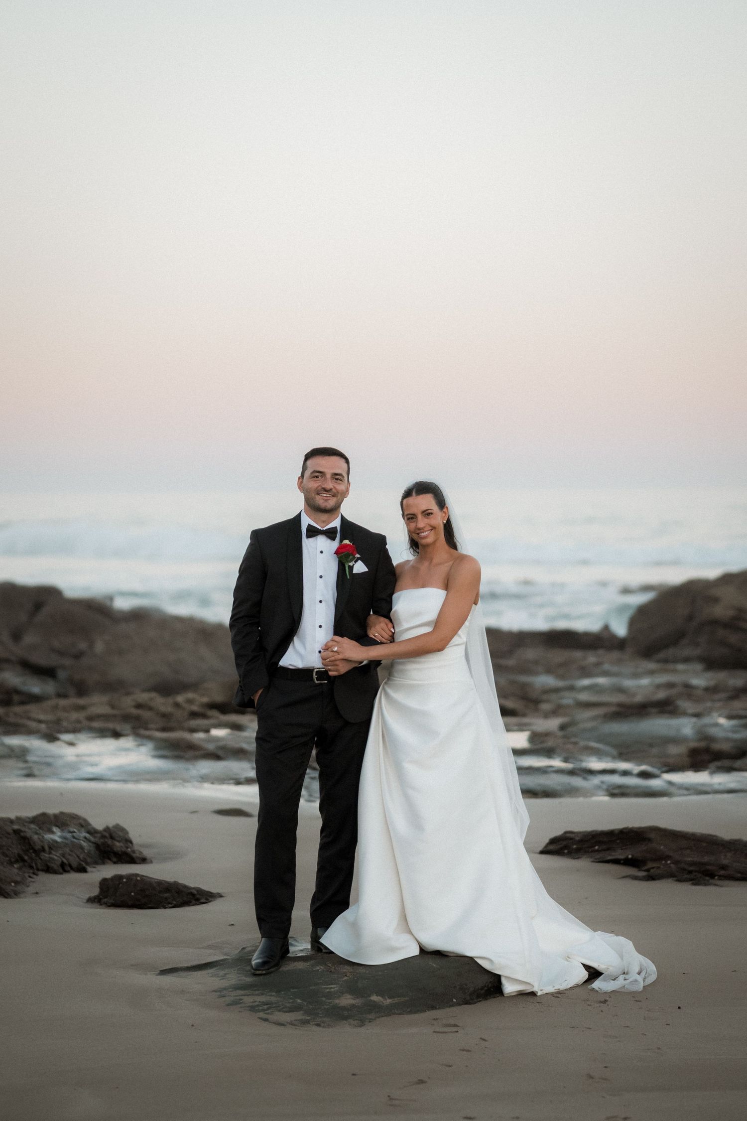 Newlyweds posing together on a rocky beach at sunset in formal wedding attire.