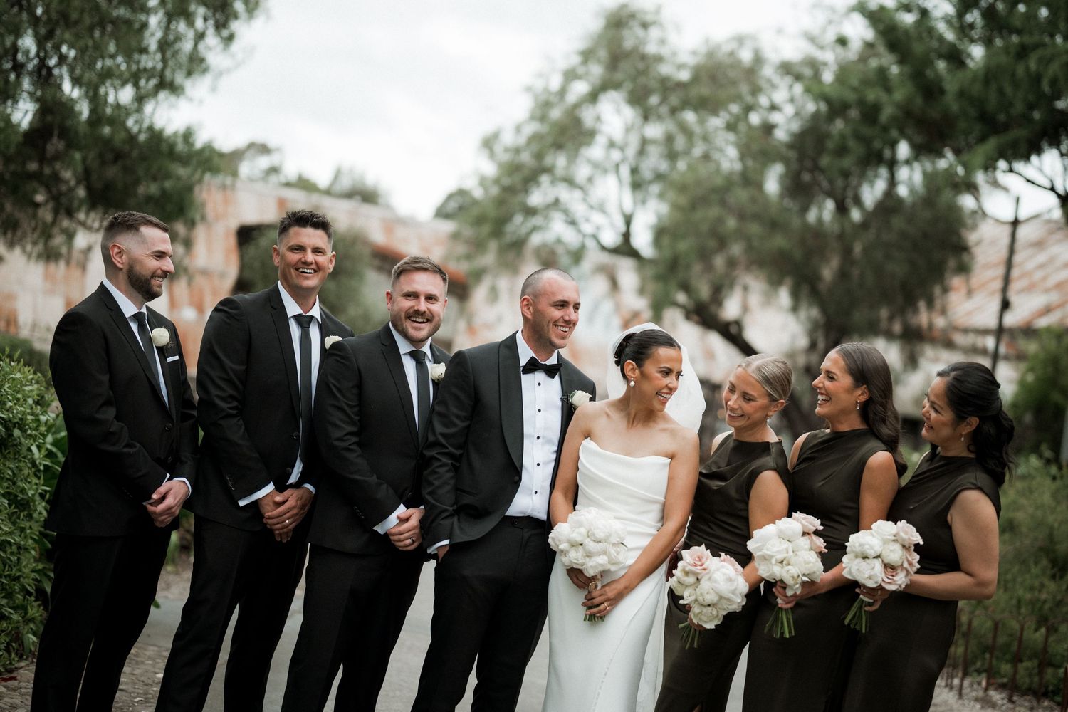 Wedding party members in formal attire pose together outdoors for group photos.