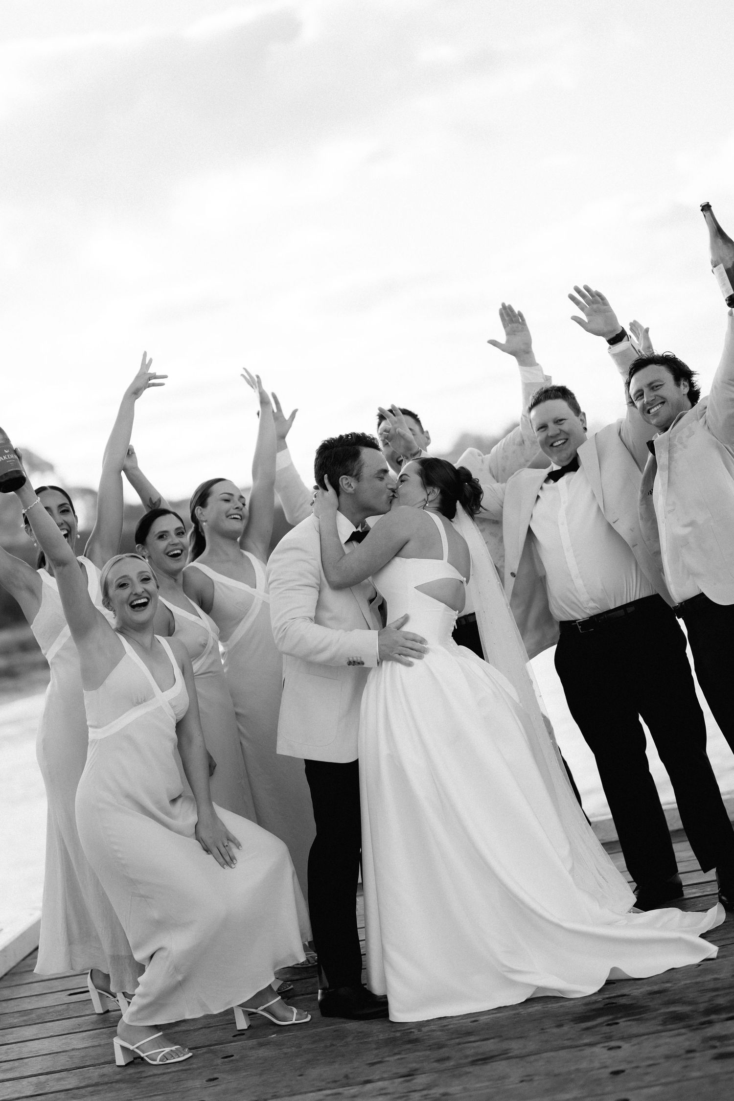 Wedding party members jump and pose playfully on a wooden deck in black and white.