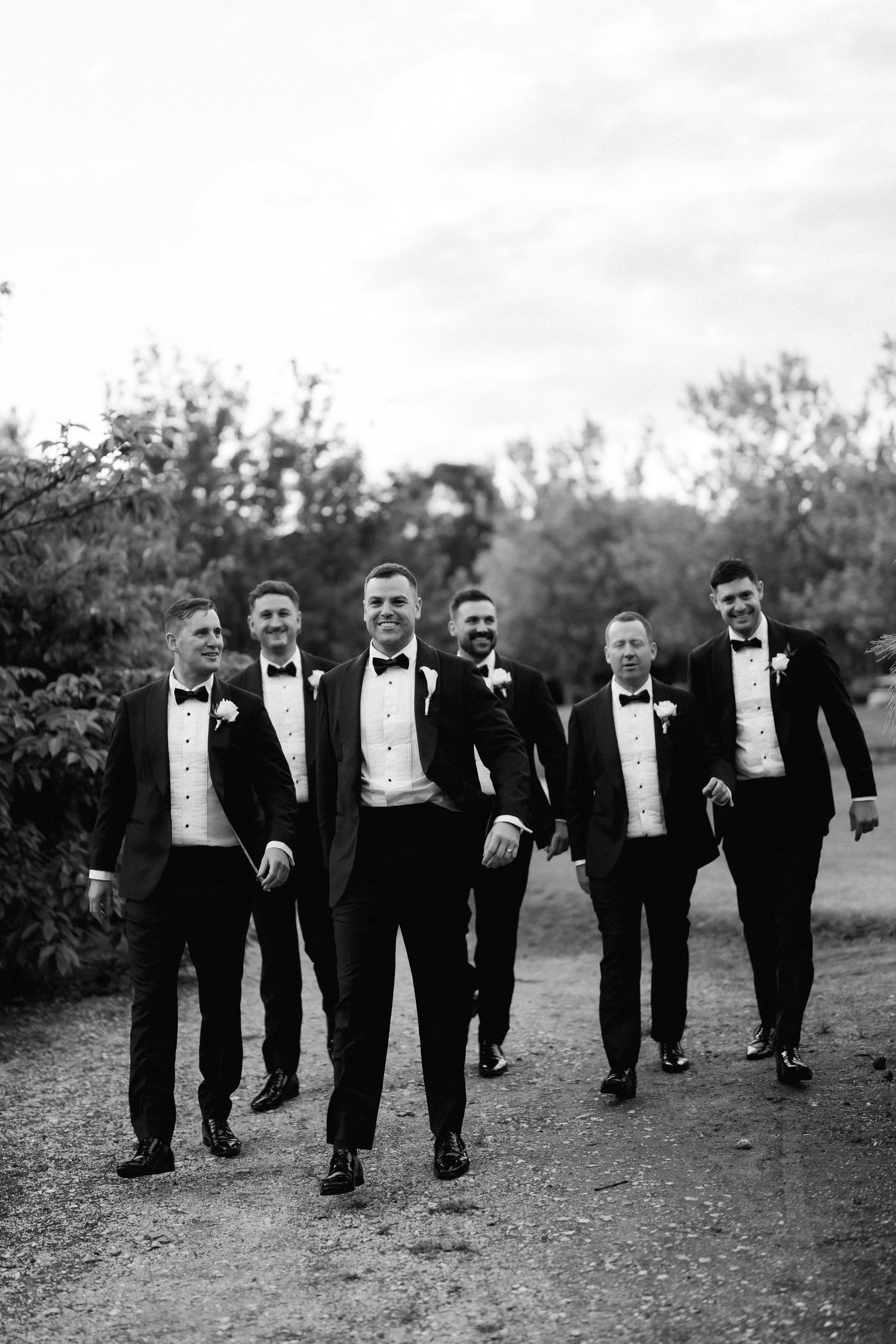 A group of groomsmen in black tuxedos and bow ties walk together outdoors in a candid black and white photo.