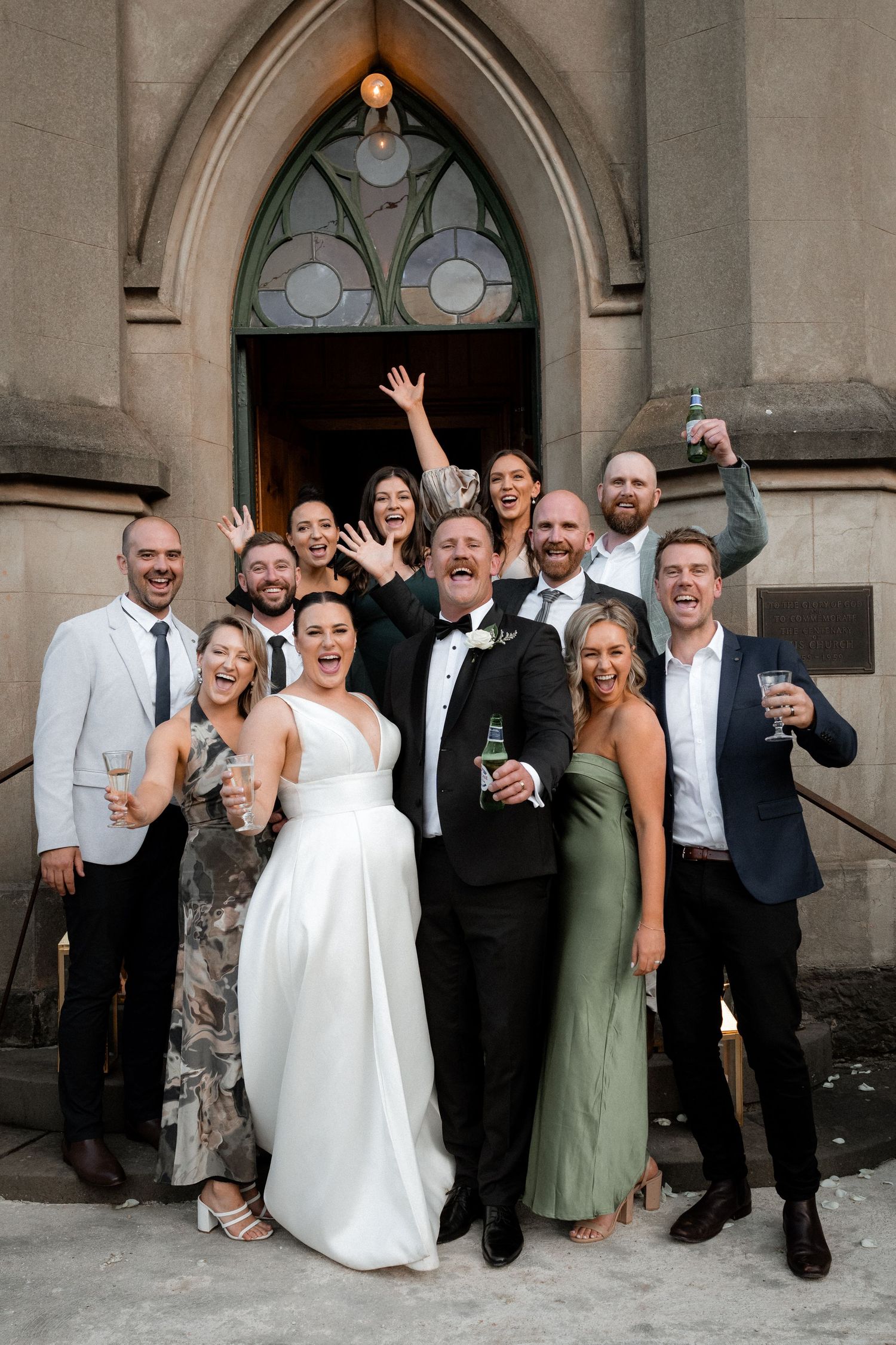 Wedding party celebrates with joyful poses and raised glasses in front of gothic church entrance doorway.