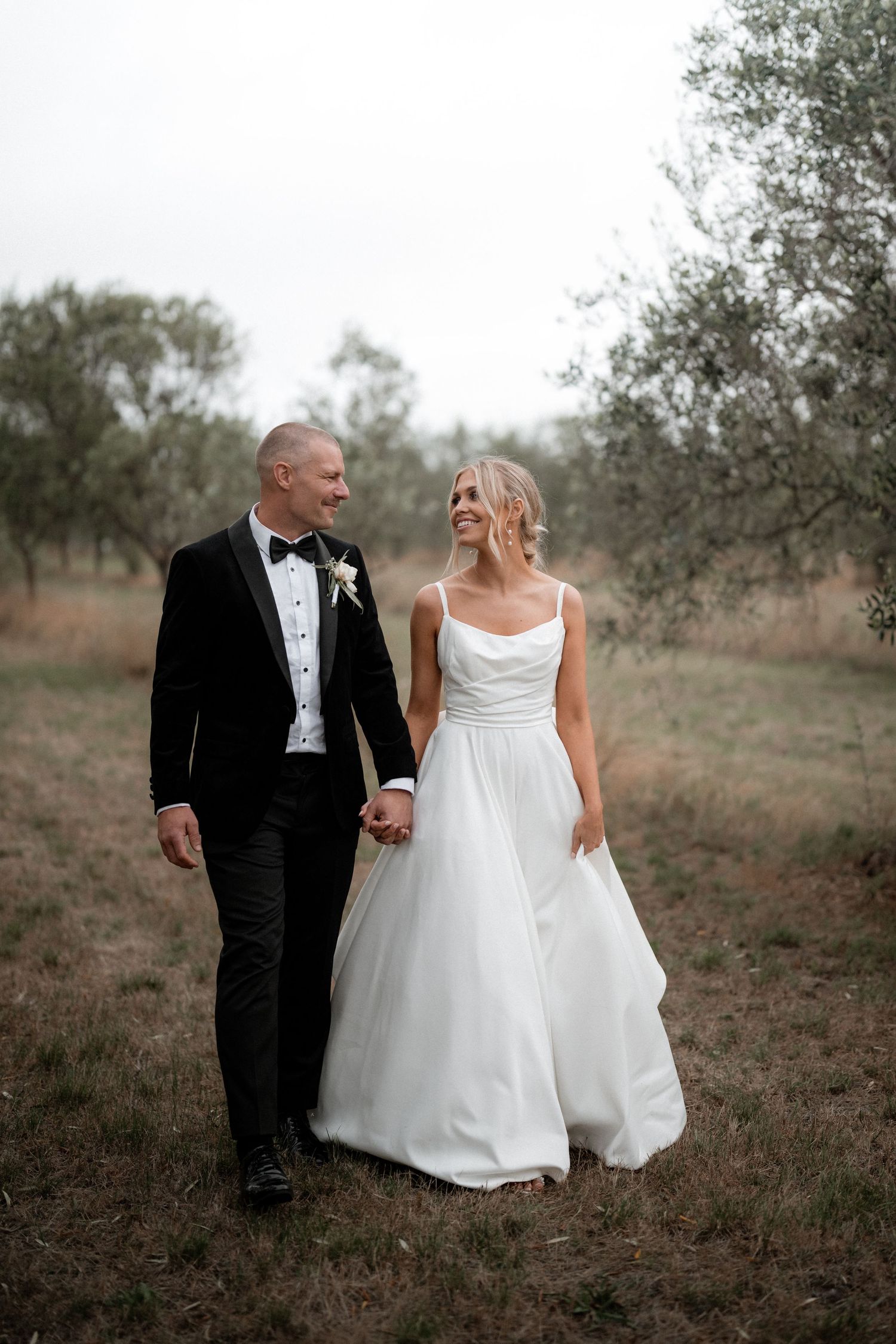 Newlyweds walking hand in hand through olive grove in formal wedding attire.