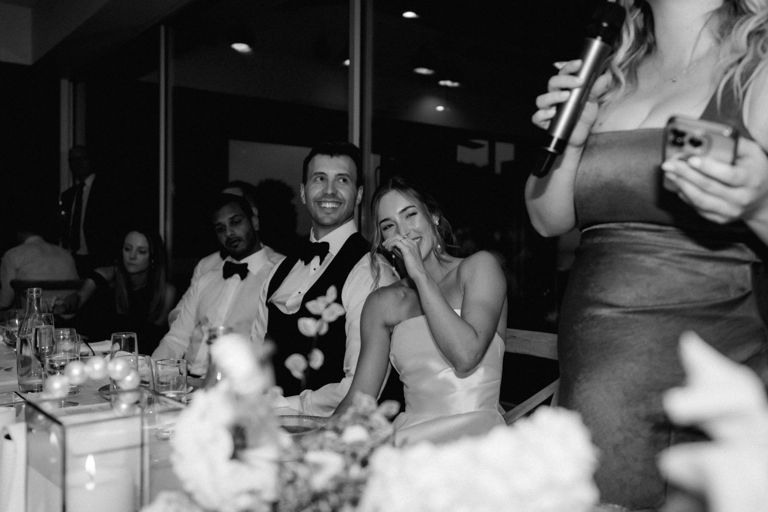 A black and white wedding reception photo showing heartfelt reactions during speeches at an elegantly set table.