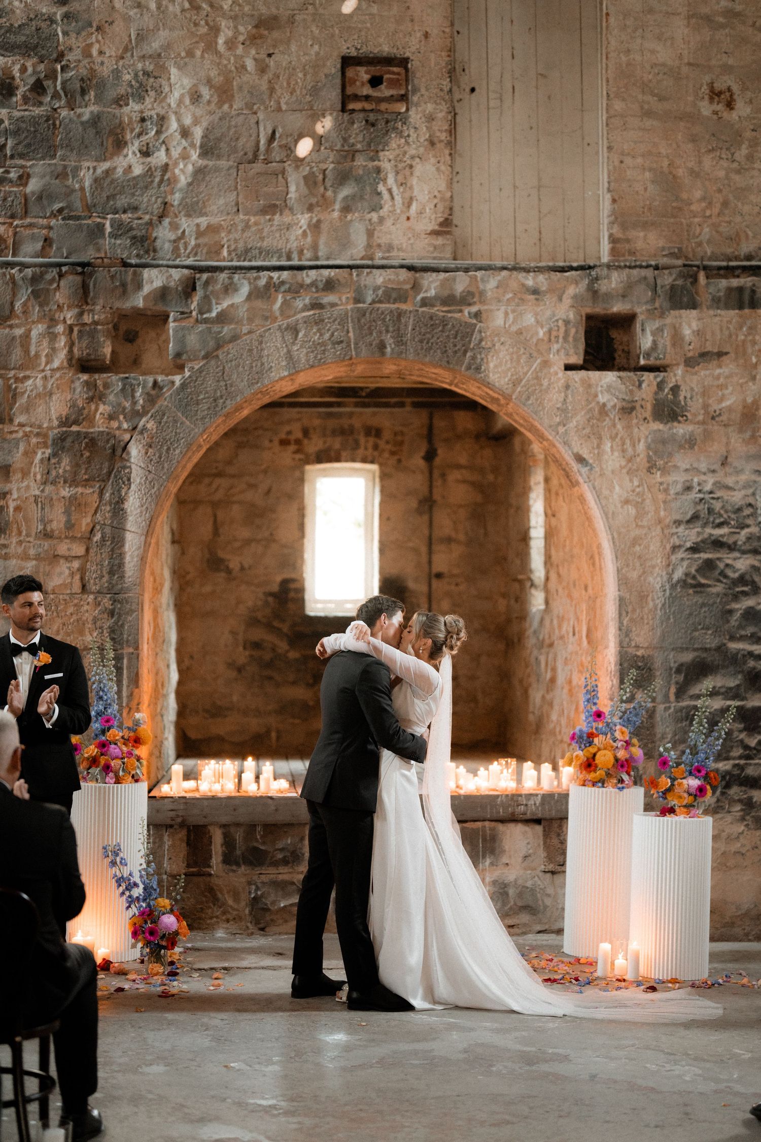 Wedding ceremony with white columns and floral arrangements in a stone-walled venue.
