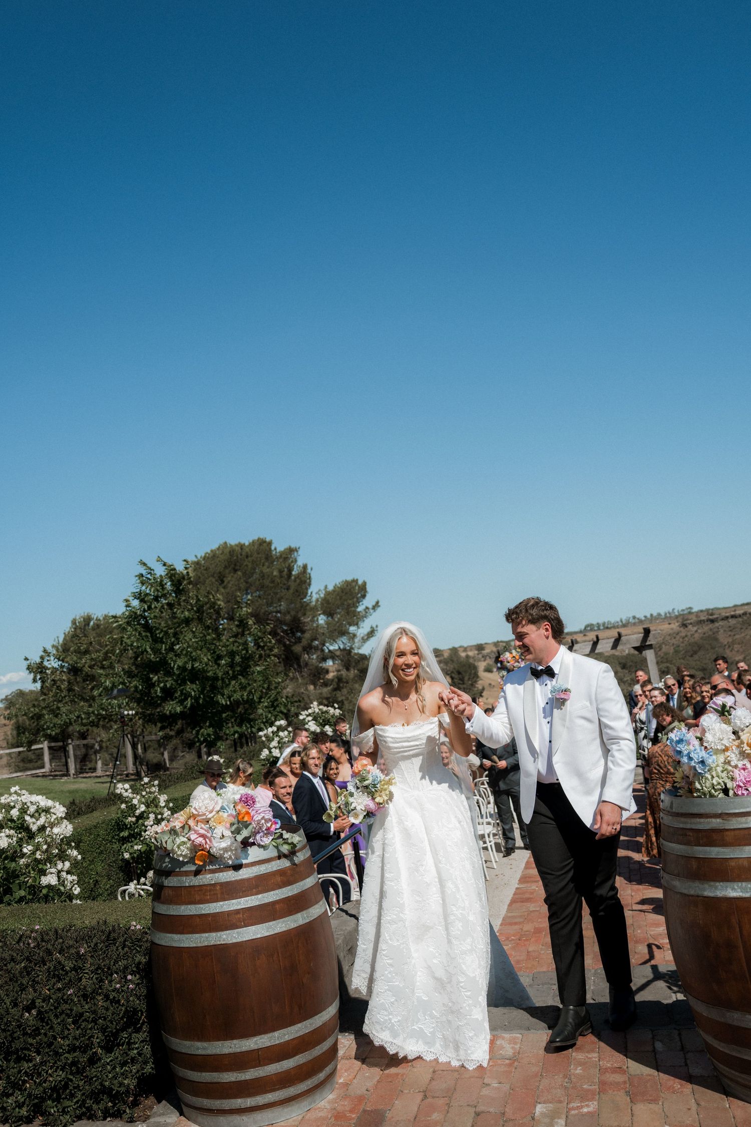 Newlyweds walk joyfully down aisle past wine barrel decorations at sunny outdoor wedding ceremony.