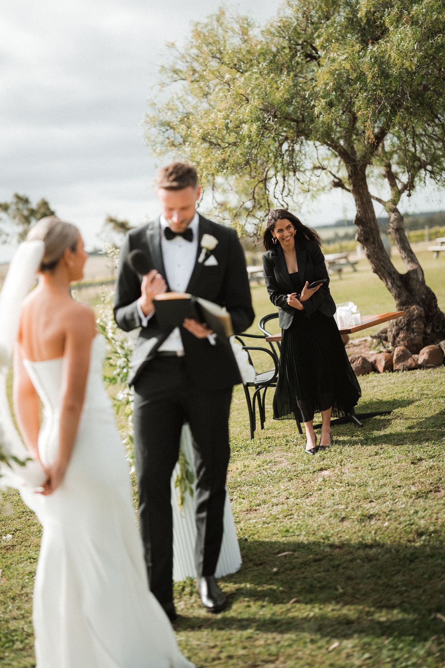 An outdoor wedding ceremony takes place under a large tree with formal attire.