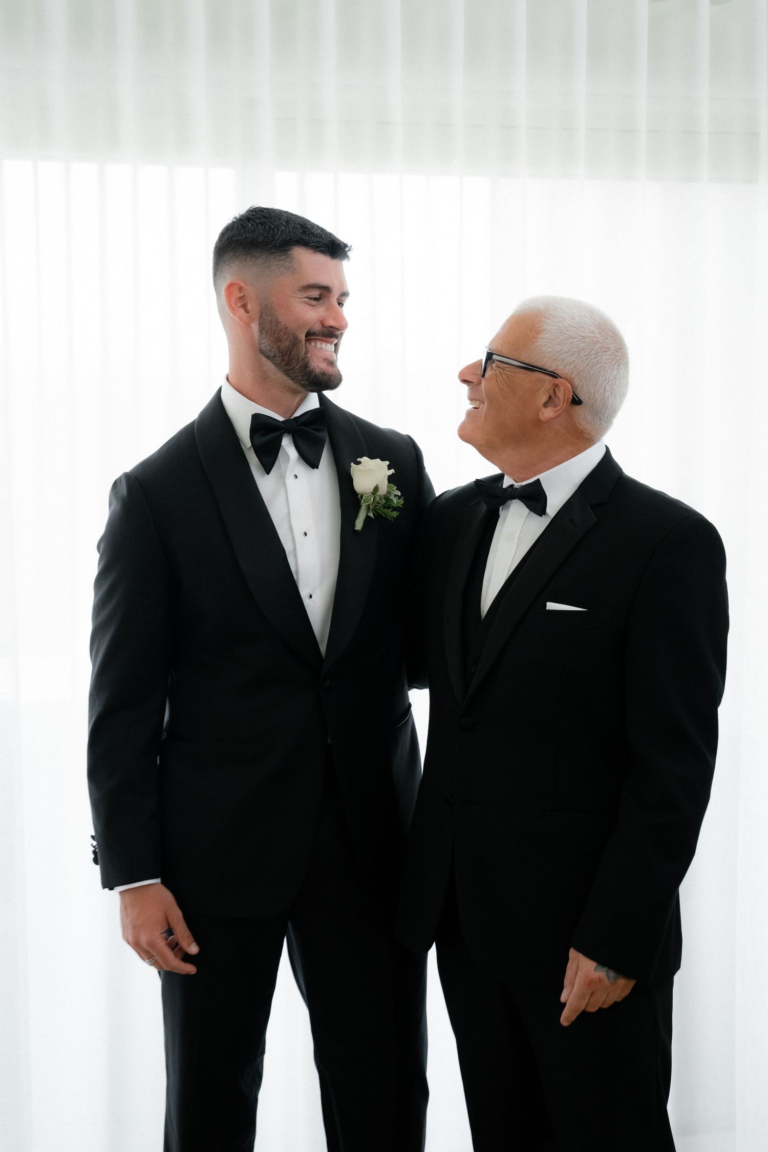 Two people in black tuxedos share a heartfelt moment against a white backdrop at a formal event.