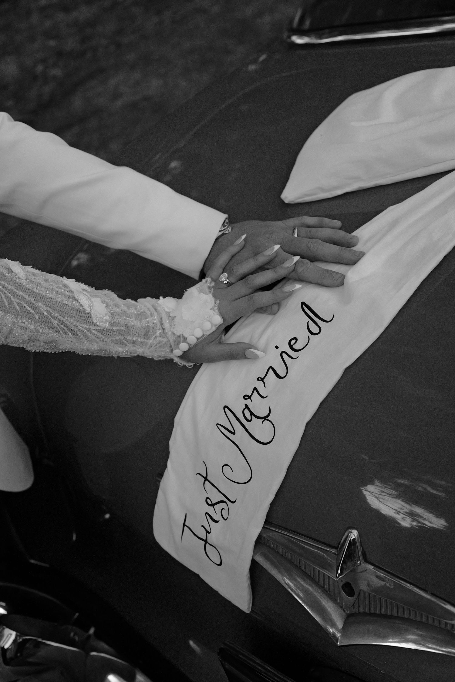Black and white photo of newlyweds hands on a Just Married banner on a vintage car hood.